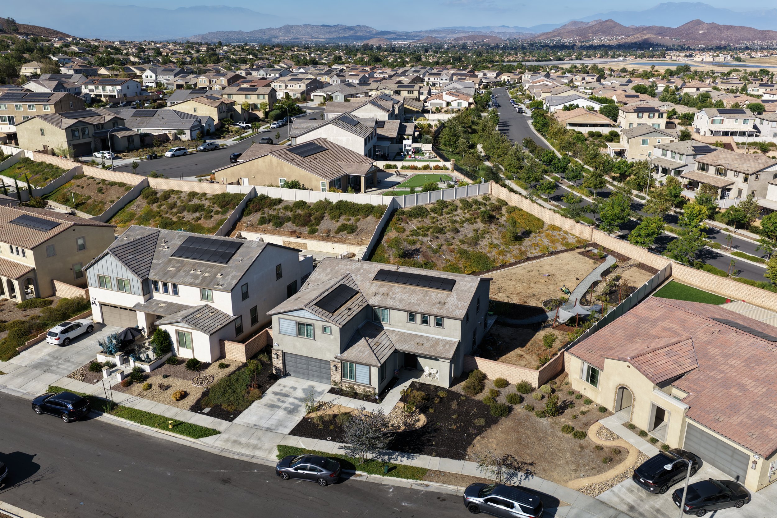 Aerial view of a suburban neighborhood with houses, some with solar panels, streets, parked cars, and a fenced backyard with a small lawn and trees. Mountains are visible in the background.