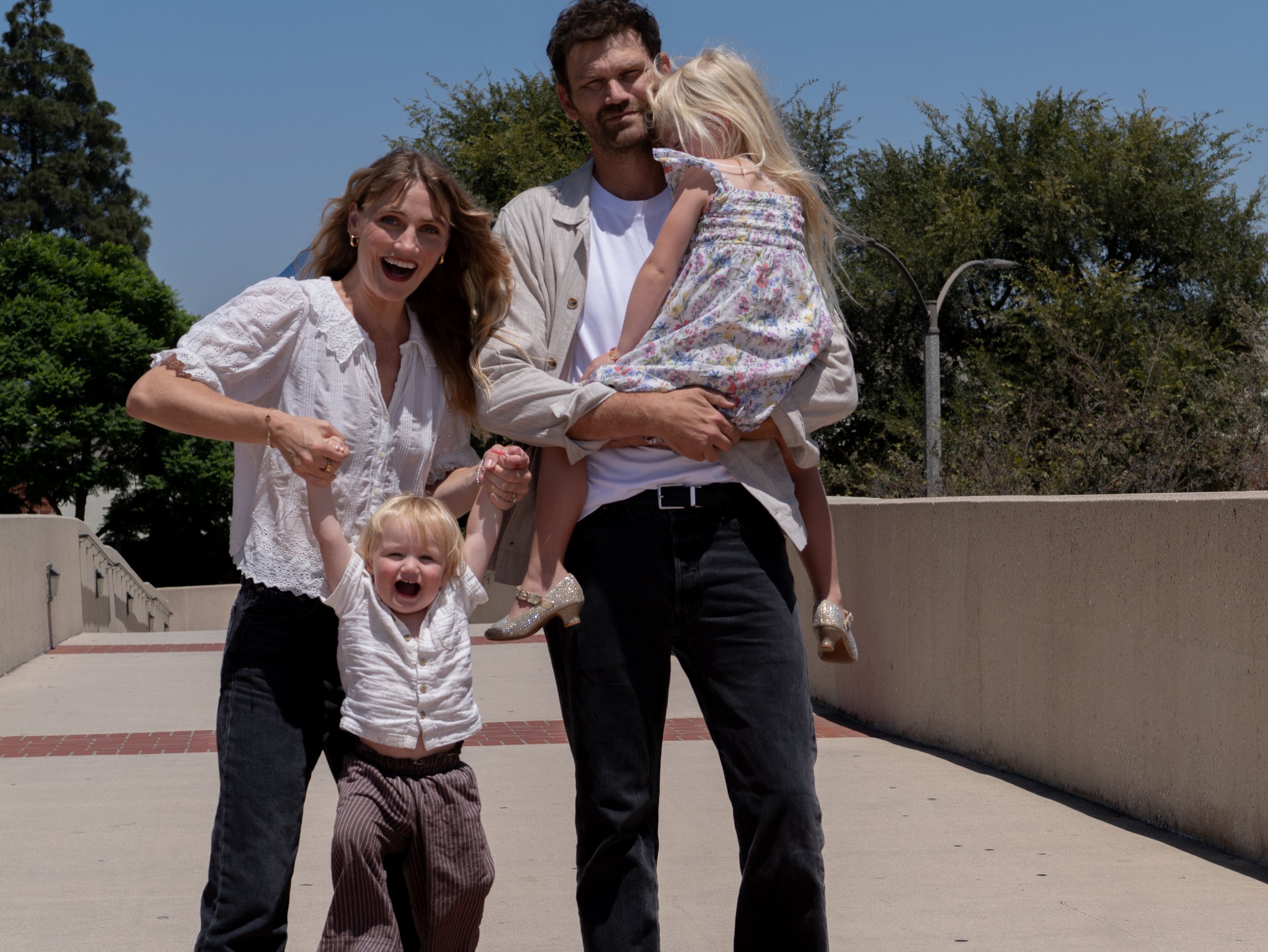 A family of four, consisting of two children, a mother, and a father, standing outdoors on a sunny day with trees and streetlights in the background. The father is holding a young girl on his shoulder, and the mother and a young boy are standing in f
