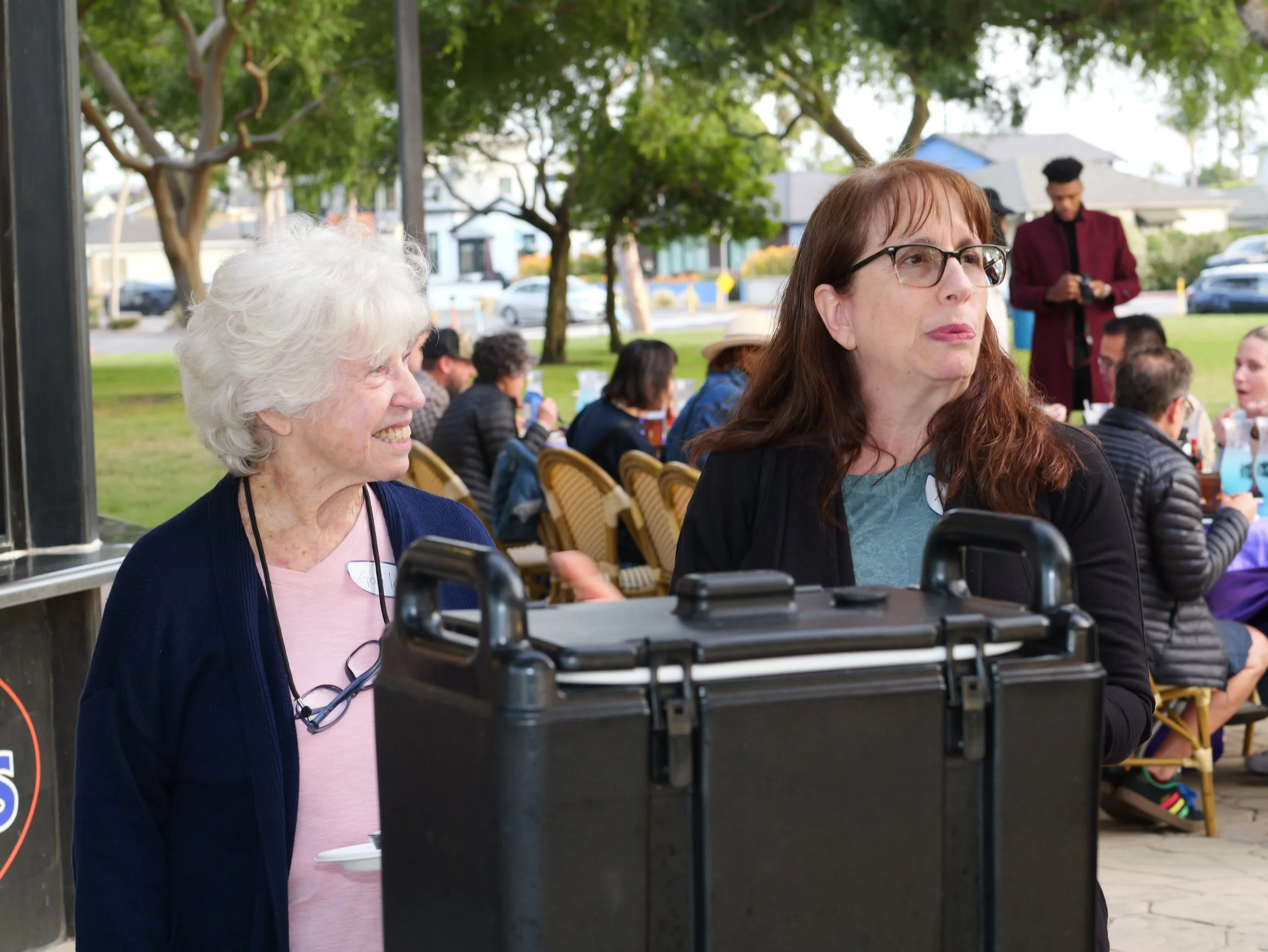 Two women sitting outdoors at a gathering, with a man standing in the background and trees overhead.