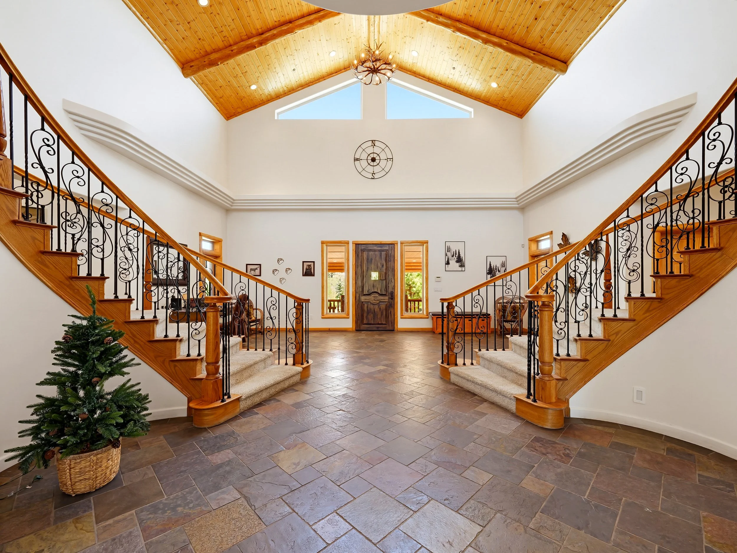 A spacious foyer with a high wooden ceiling and a modern chandelier. A staircase with decorative black wrought iron railings leads to an upper level. A small Christmas tree in a basket is placed on the left. The front door is wooden with side panels, flanked by windows. The floor is tiled with multicolored stone tiles.