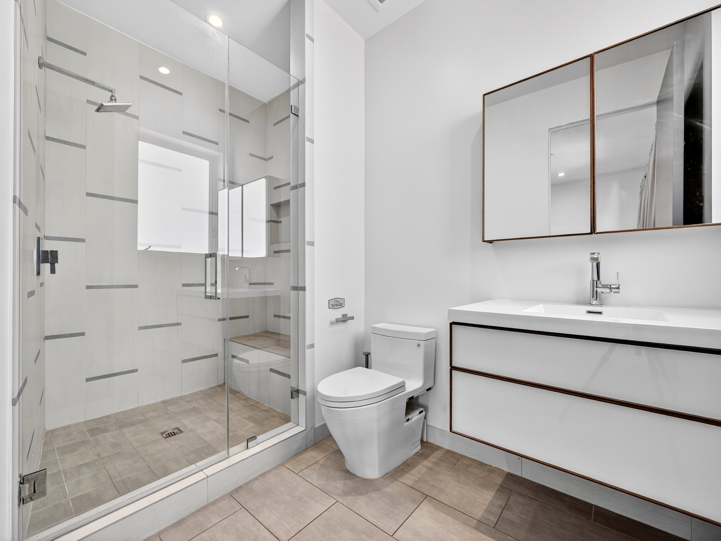 Modern bathroom with a walk-in glass shower, white toilet, and white vanity with brown trim. The shower has sliding glass doors with a showerhead, and the sink has a sleek faucet. The walls are white, and the floor is beige tile.