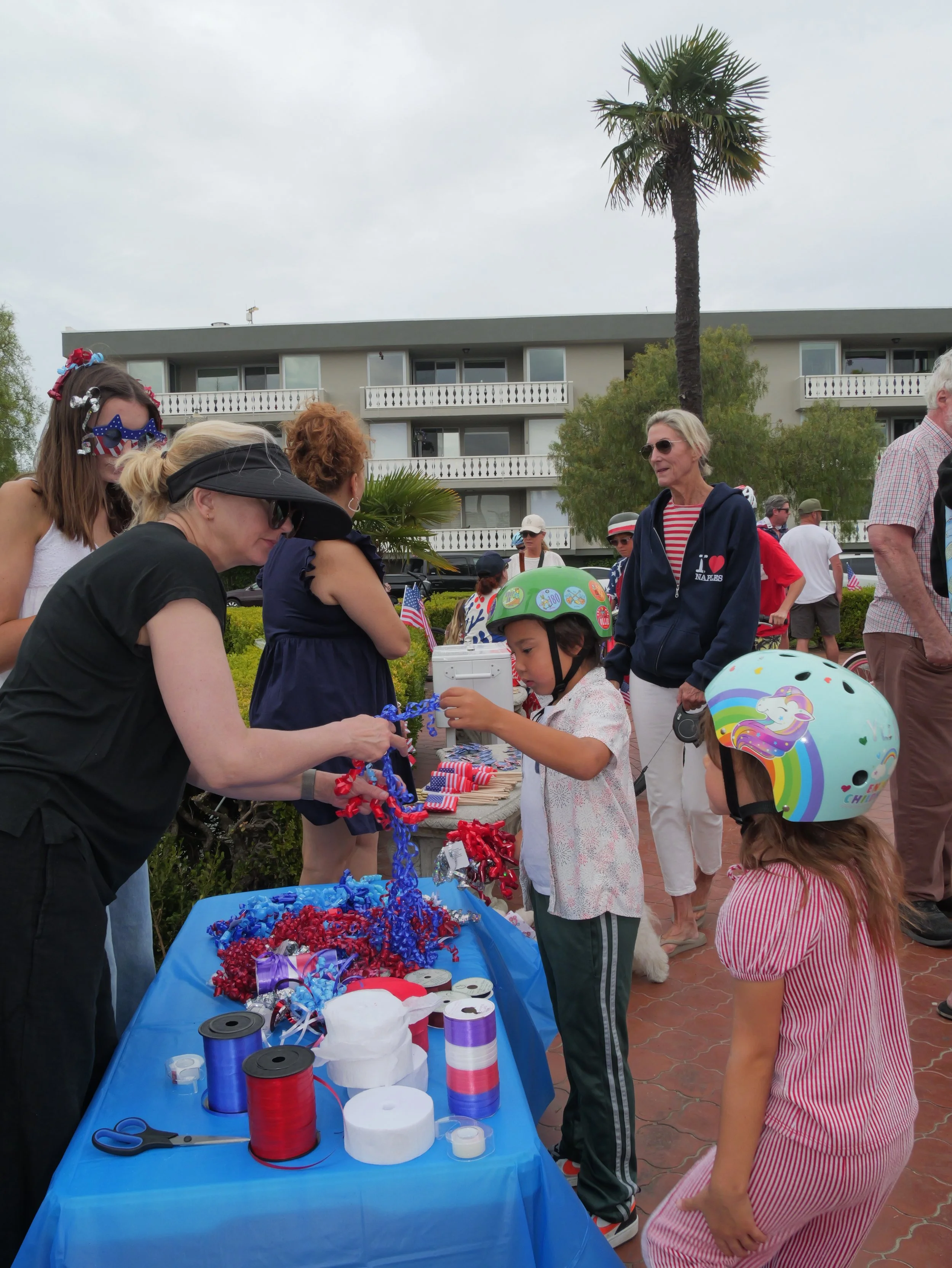 People gather around a table outdoors with patriotic decorations, ribbons, and craft supplies, likely for a celebration or community event, with apartment buildings, a large palm tree, and cloudy sky in the background.