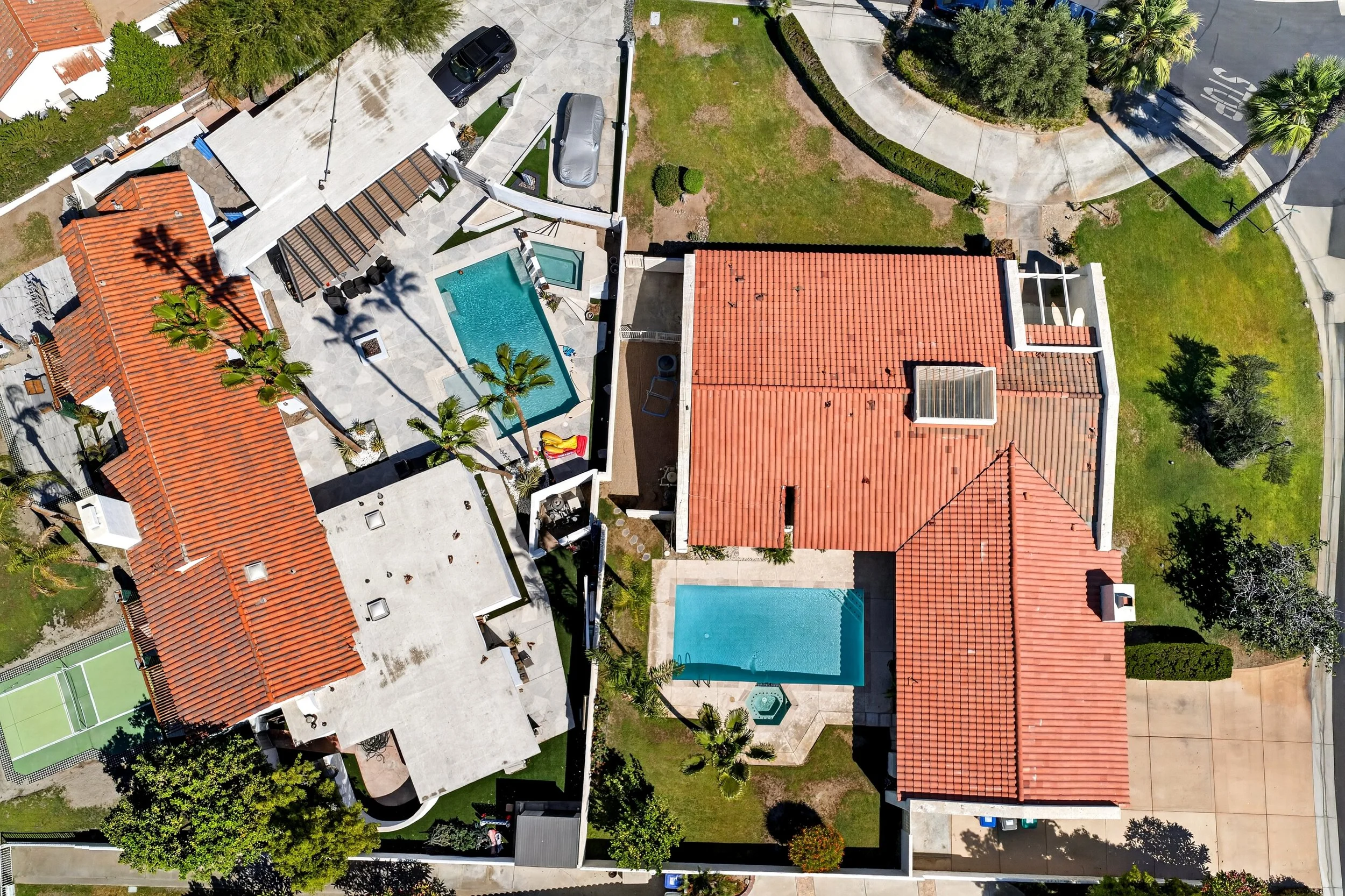 Aerial view of residential backyard with swimming pool, patio area, palm trees, and neighboring houses with red-tiled roofs.