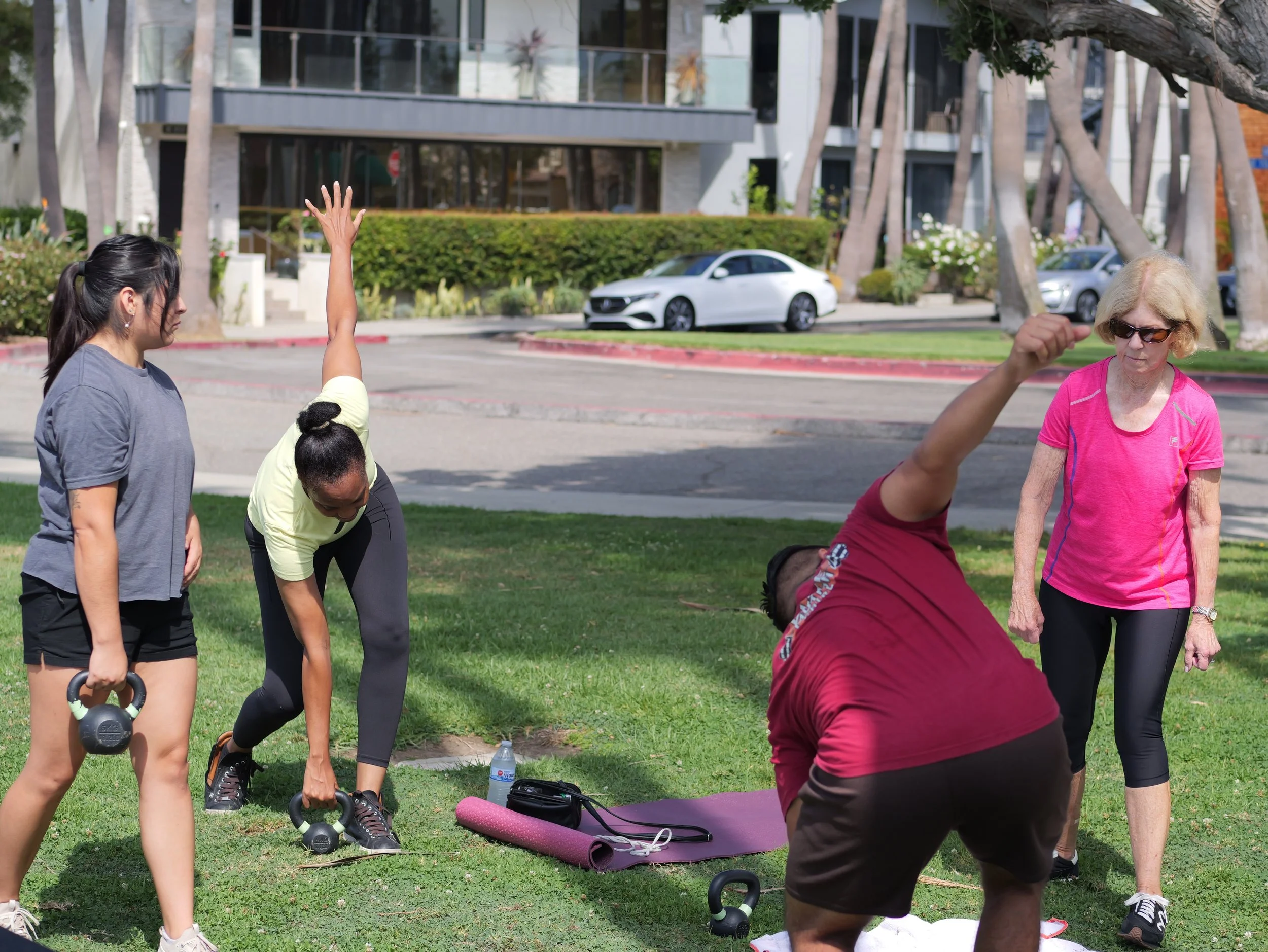 A group of women exercising outdoors in a park on a sunny day, with some holding kettlebells and others standing around a yoga mat.
