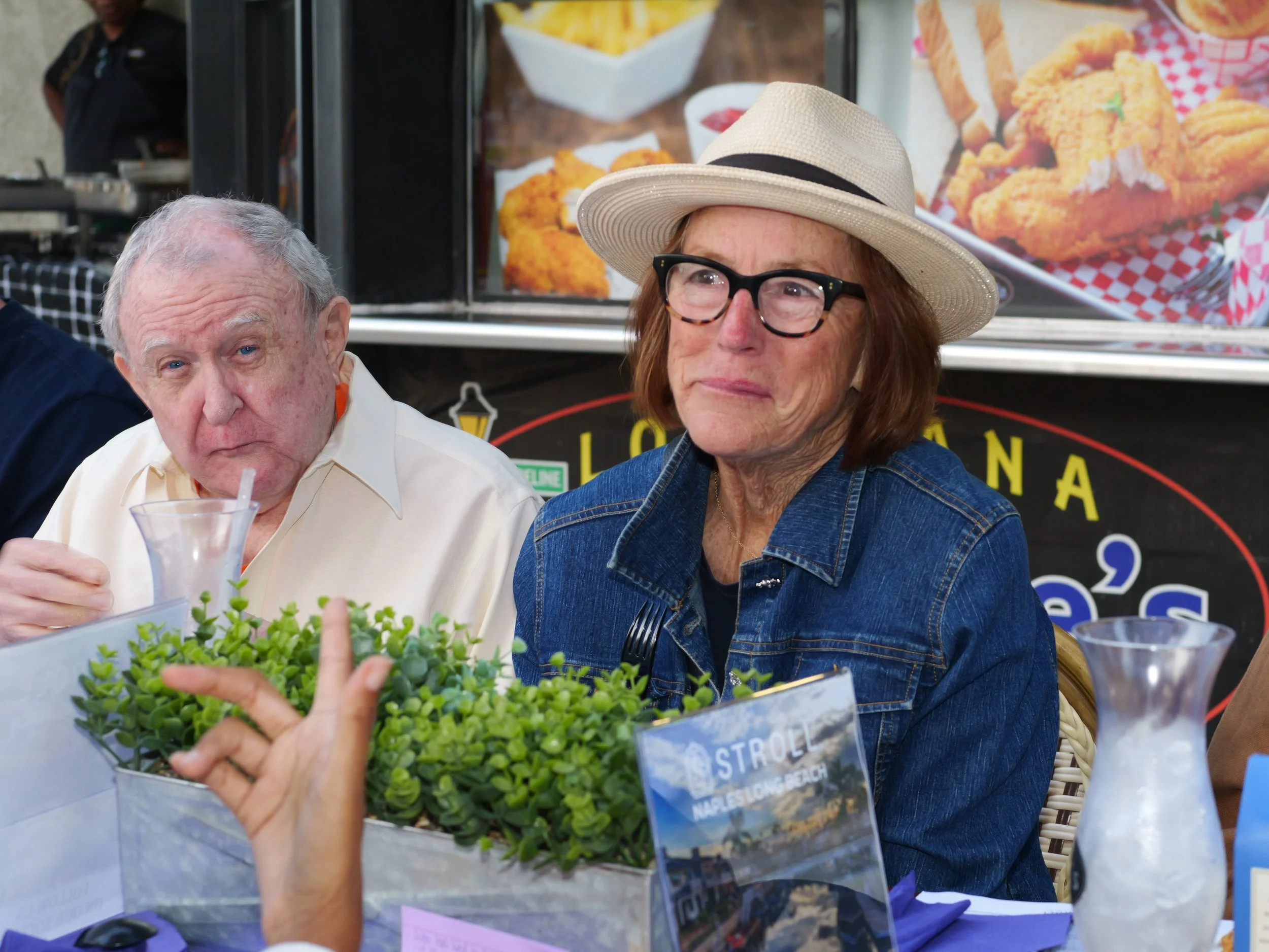 A woman wearing glasses and a wide-brimmed hat looks at a container of green succulents at an outdoor food stand, with a blurred background of fried foods and fries.