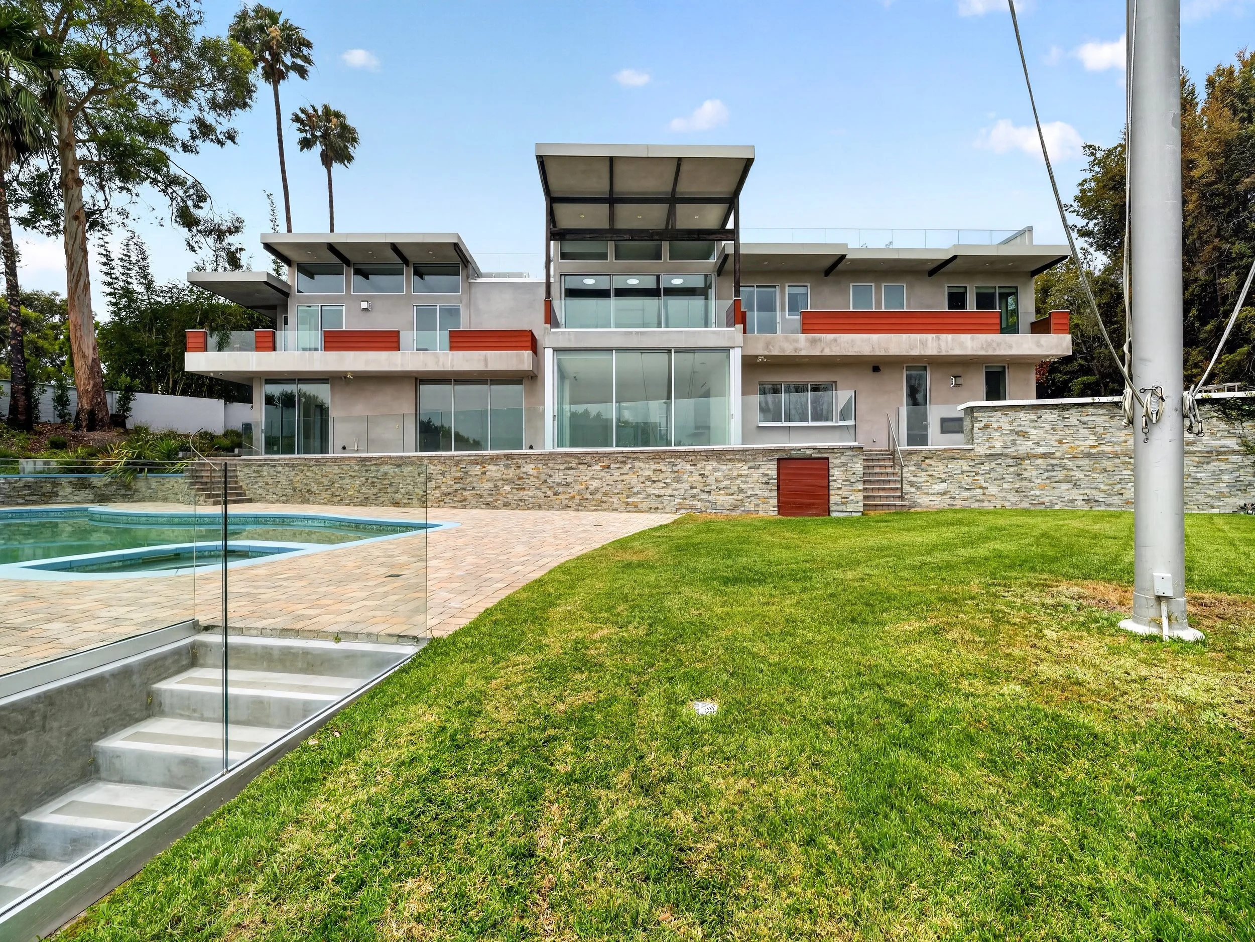 Modern multi-story house with large glass windows, balconies with red accents, and a swimming pool in the foreground, surrounded by green lawn and tall trees under a partly cloudy sky.
