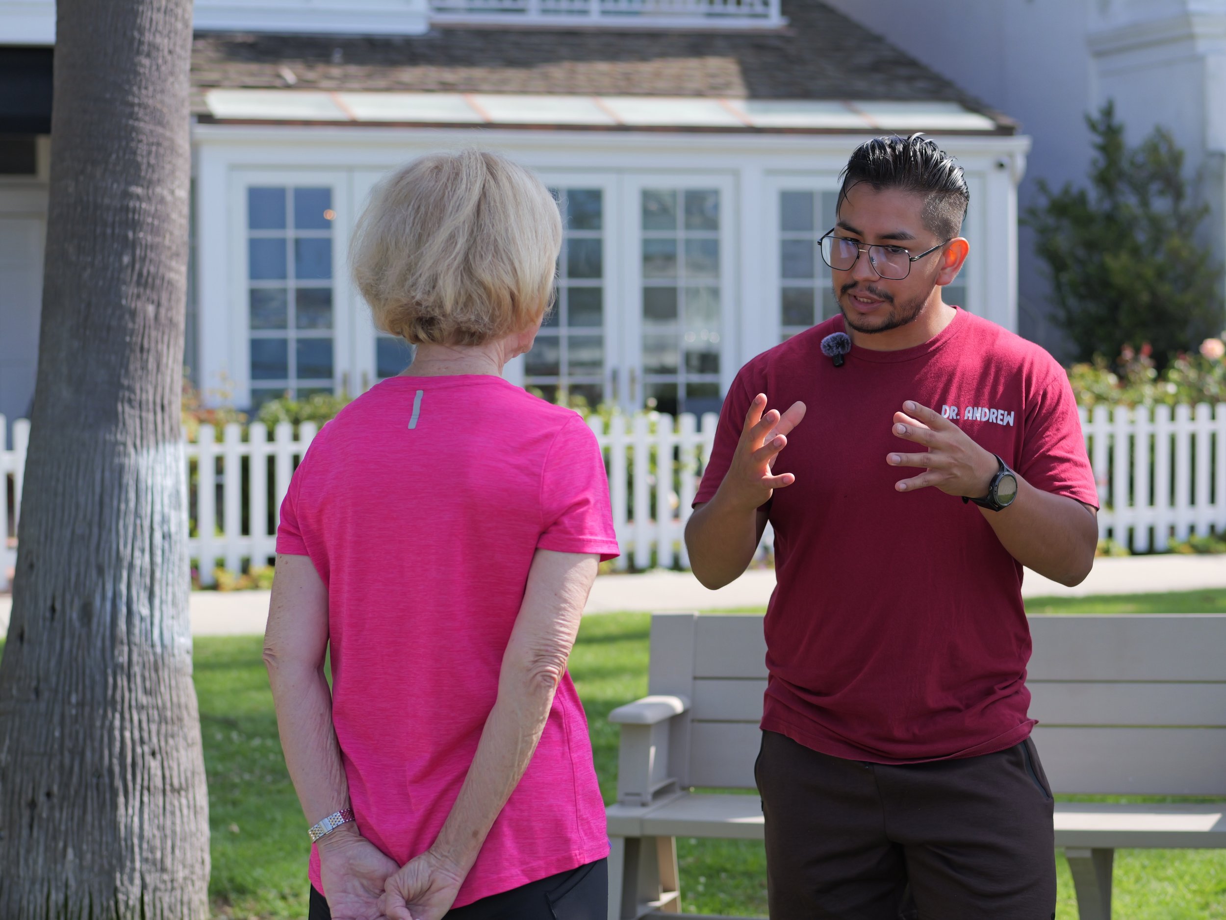 A male healthcare professional speaking to an older woman outdoors in front of a house with a white fence, green lawn, and trees.