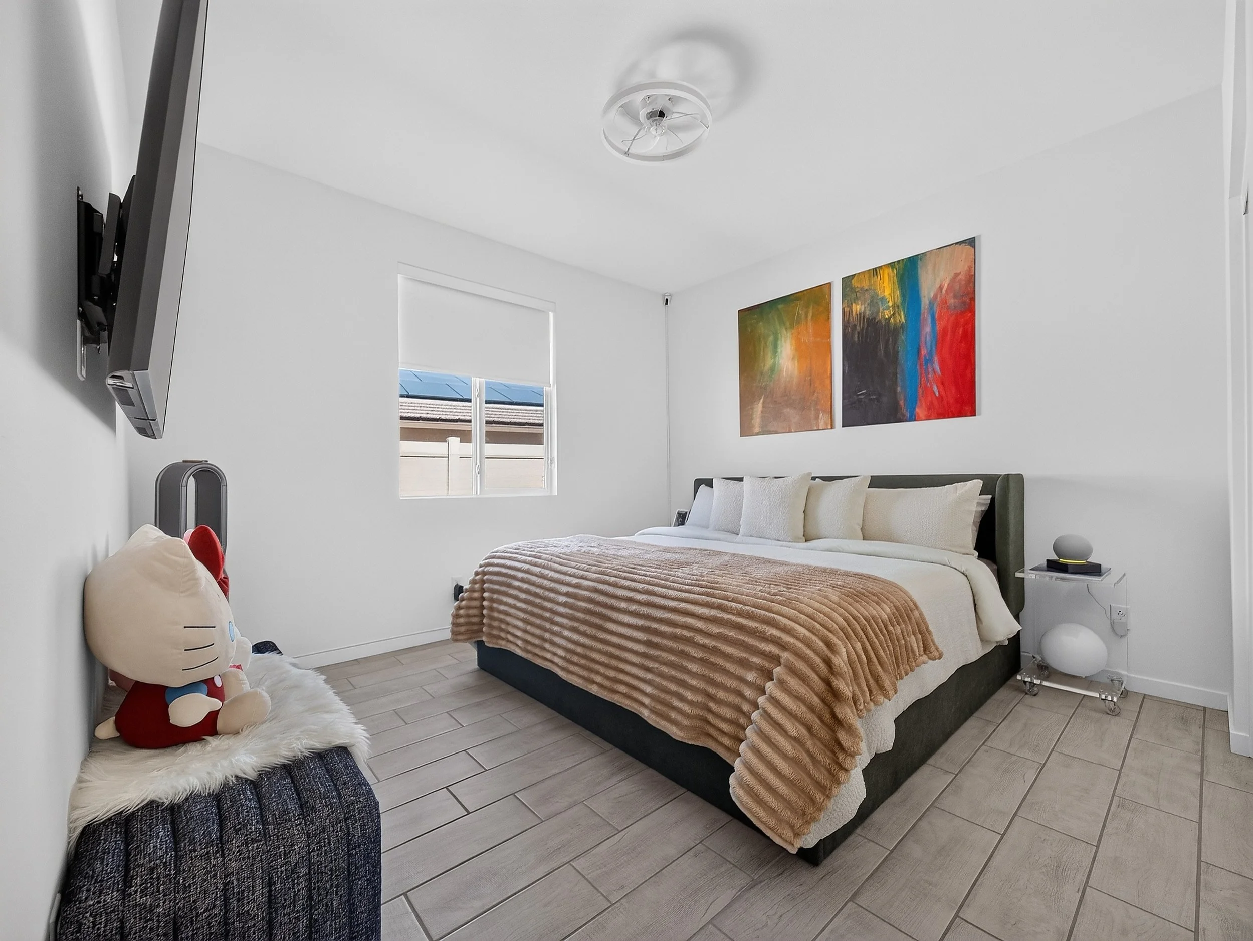 A bedroom with a bed covered in a beige textured blanket and white pillows. Two colorful abstract paintings hang on the white wall above the bed. There is a window with a white blind, and a ceiling fan. A stuffed Hello Kitty toy and a plush white rug