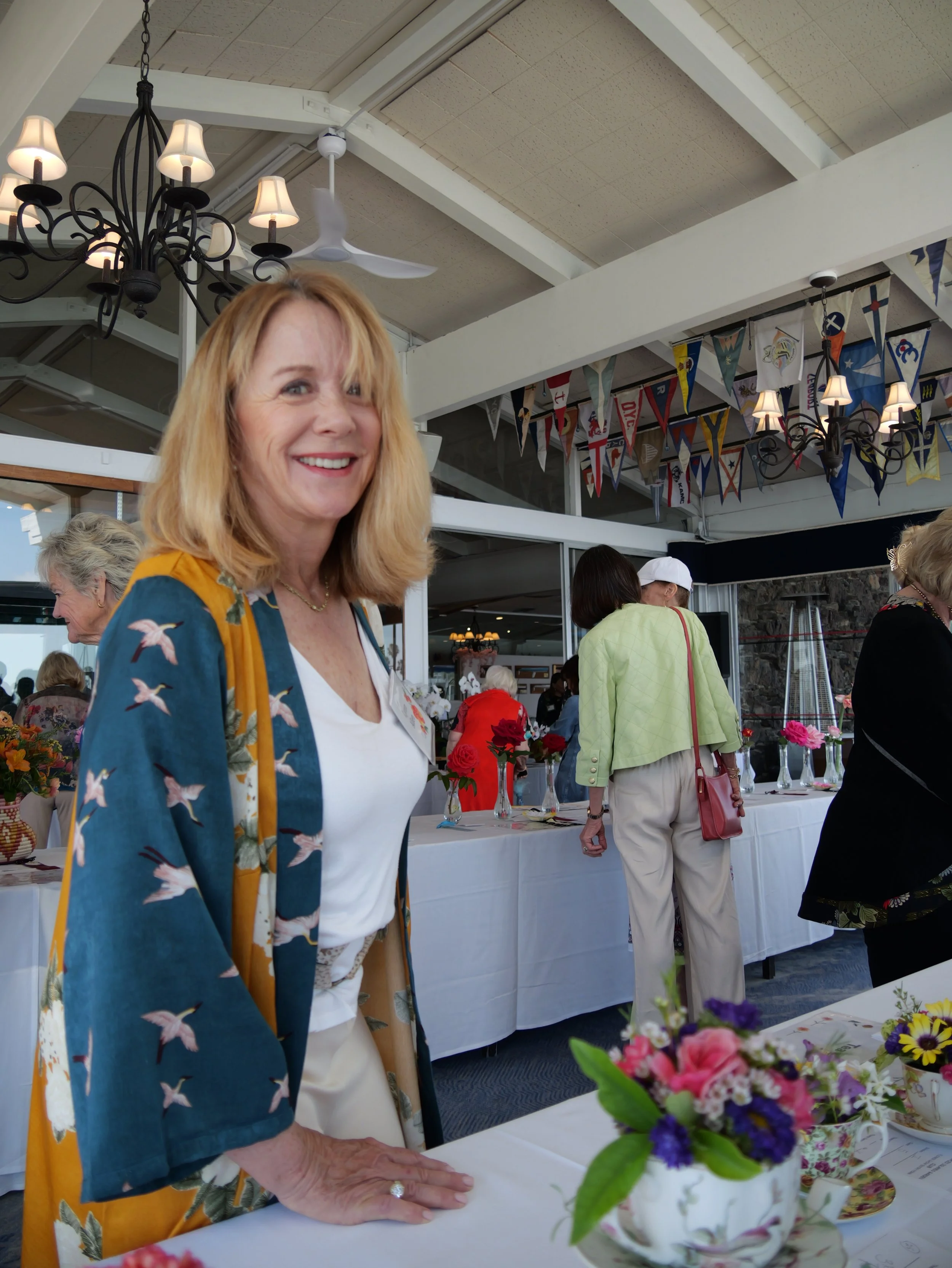 A woman with blonde hair and a patterned jacket smiling at a social gathering, with colorful flags hanging above and tables with flowers in the background.