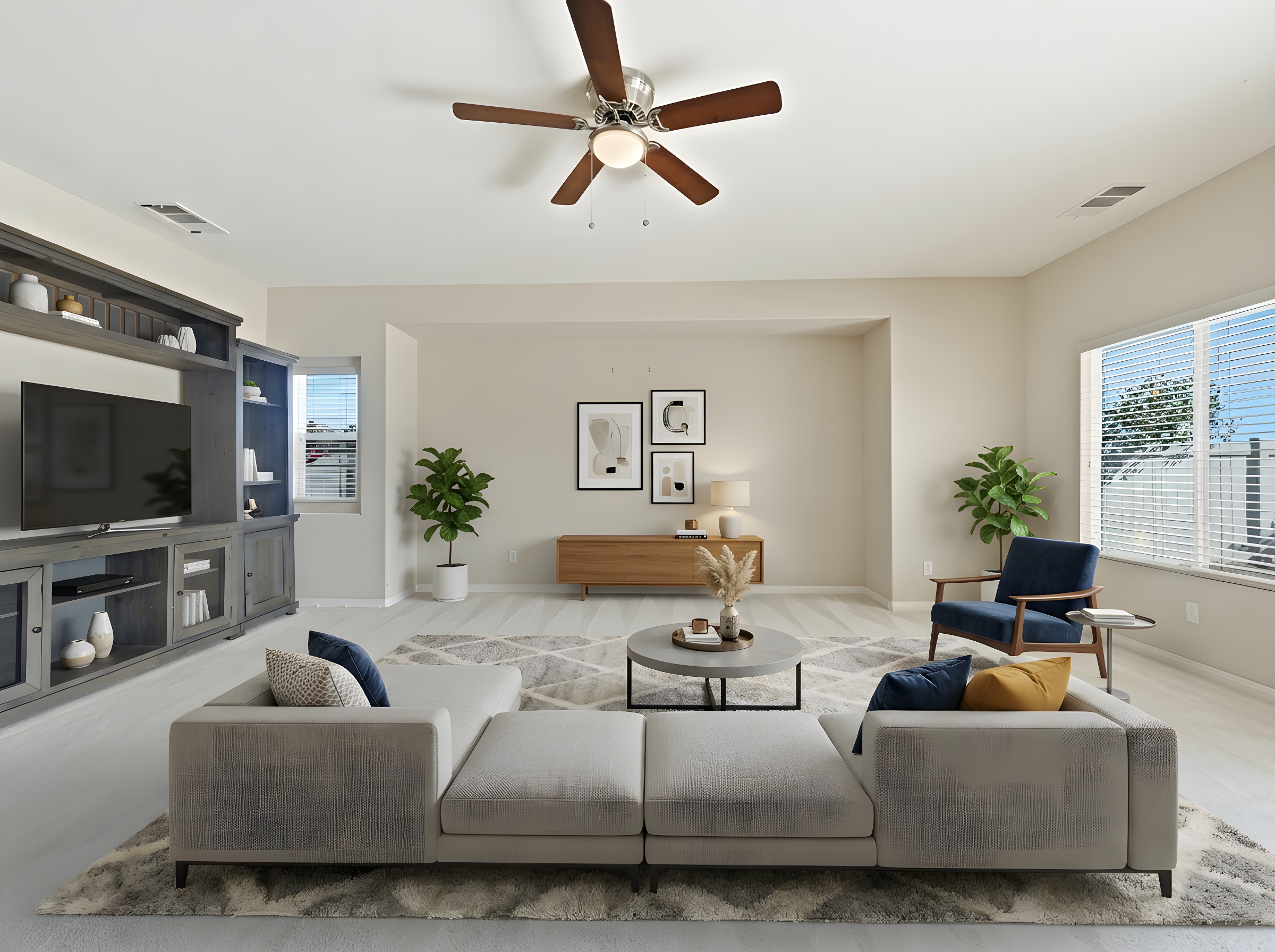 Modern living room with white walls, large windows with blinds, and a ceiling fan with wooden blades. The room has a gray entertainment center, framed abstract art, green potted plants, a beige sofa with pillows, a coffee table with decorations, and a dark blue armchair.