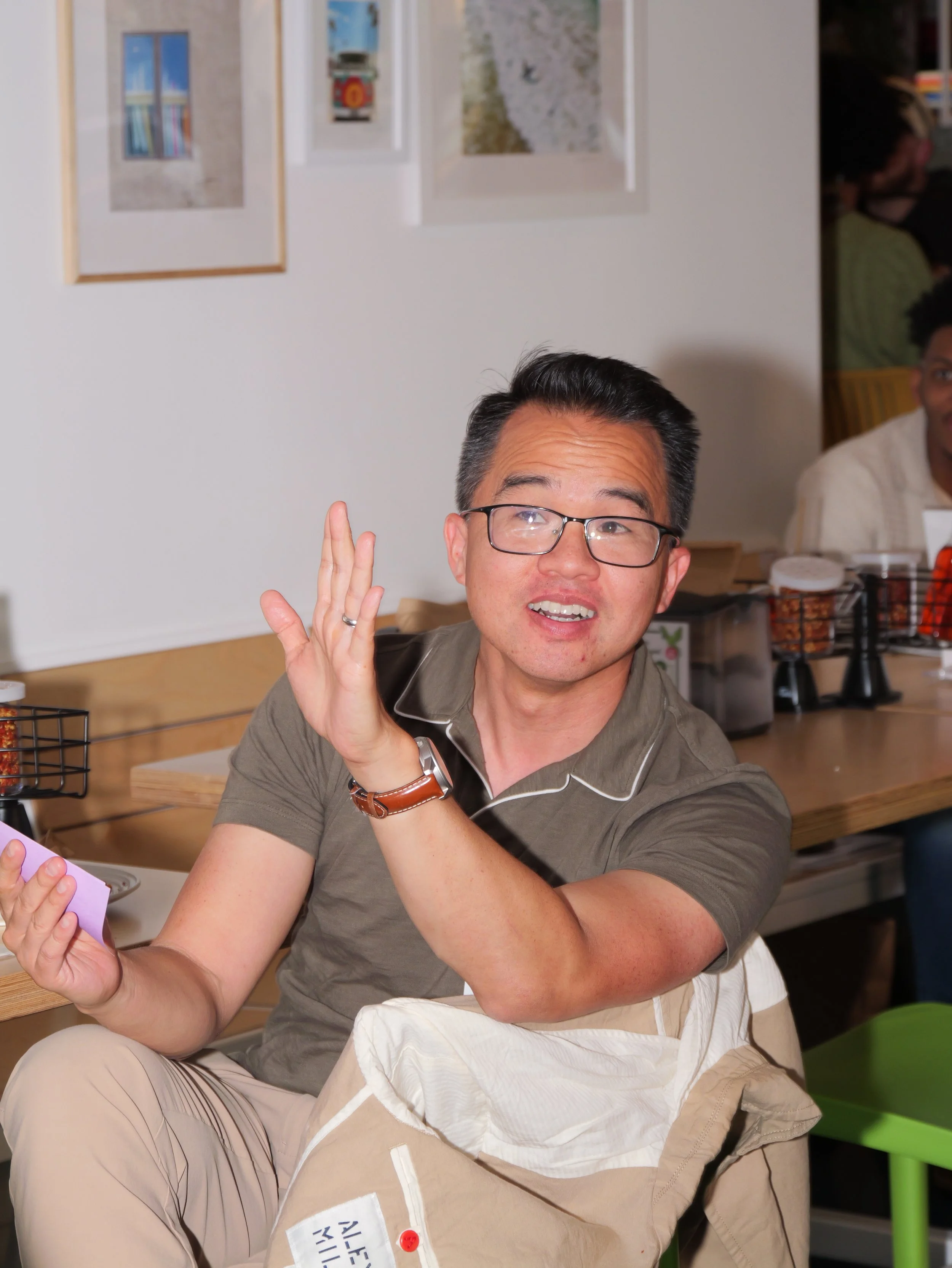 A man wearing glasses and a watch seated at a table, raising his hand, with other people in the background at a cafe or restaurant.