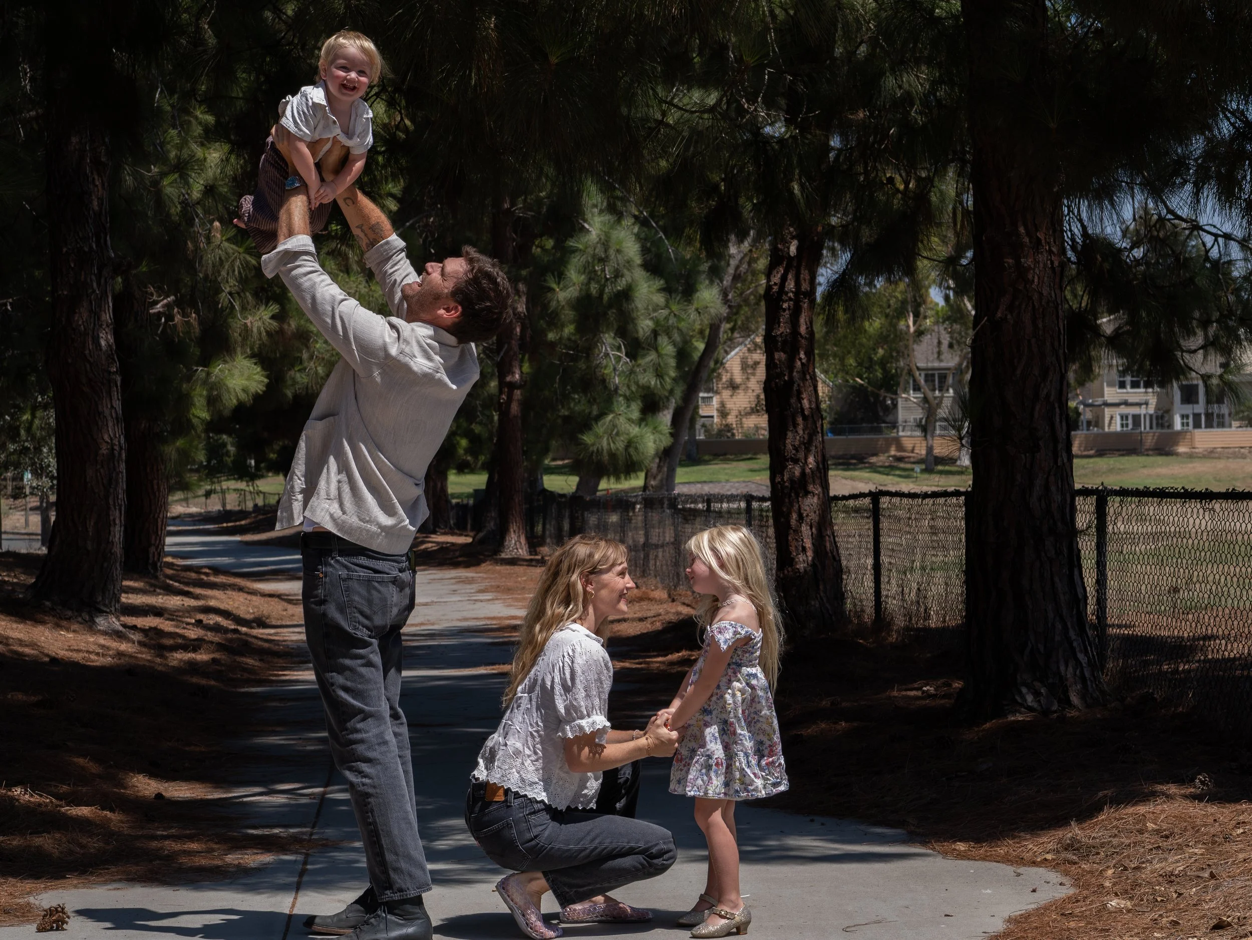 A man is kneeling on one knee, holding hands with a young girl, while another girl stands nearby, holding their hands. The man is lifting a young child into the air, and they are all outdoors among trees, with some buildings visible in the background