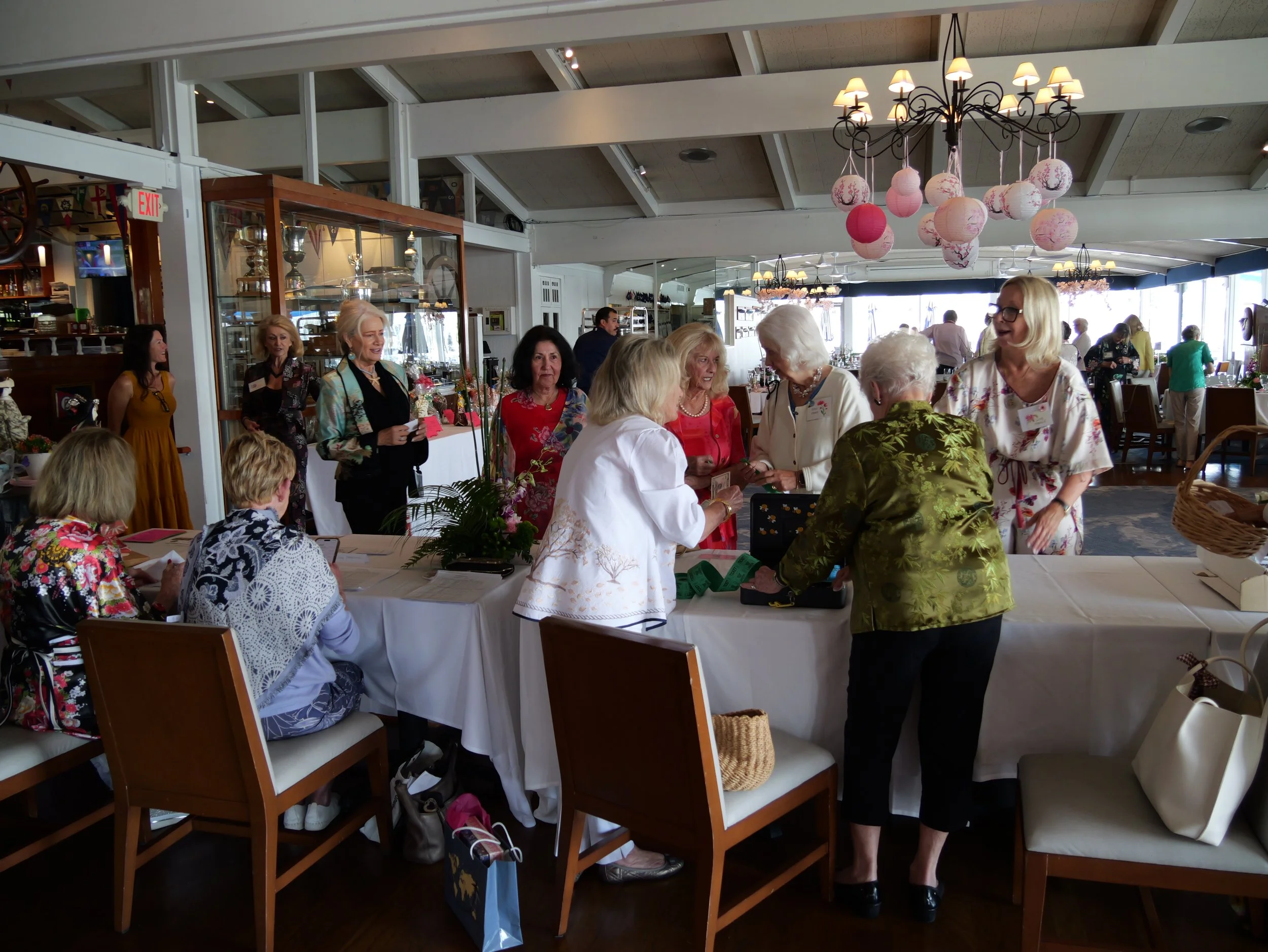 A group of mostly elderly women gathered around a long table inside a restaurant or event space, engaging in conversation. The room has hanging paper lanterns and chandeliers, large windows, and display cases with trophies and kitchenware.