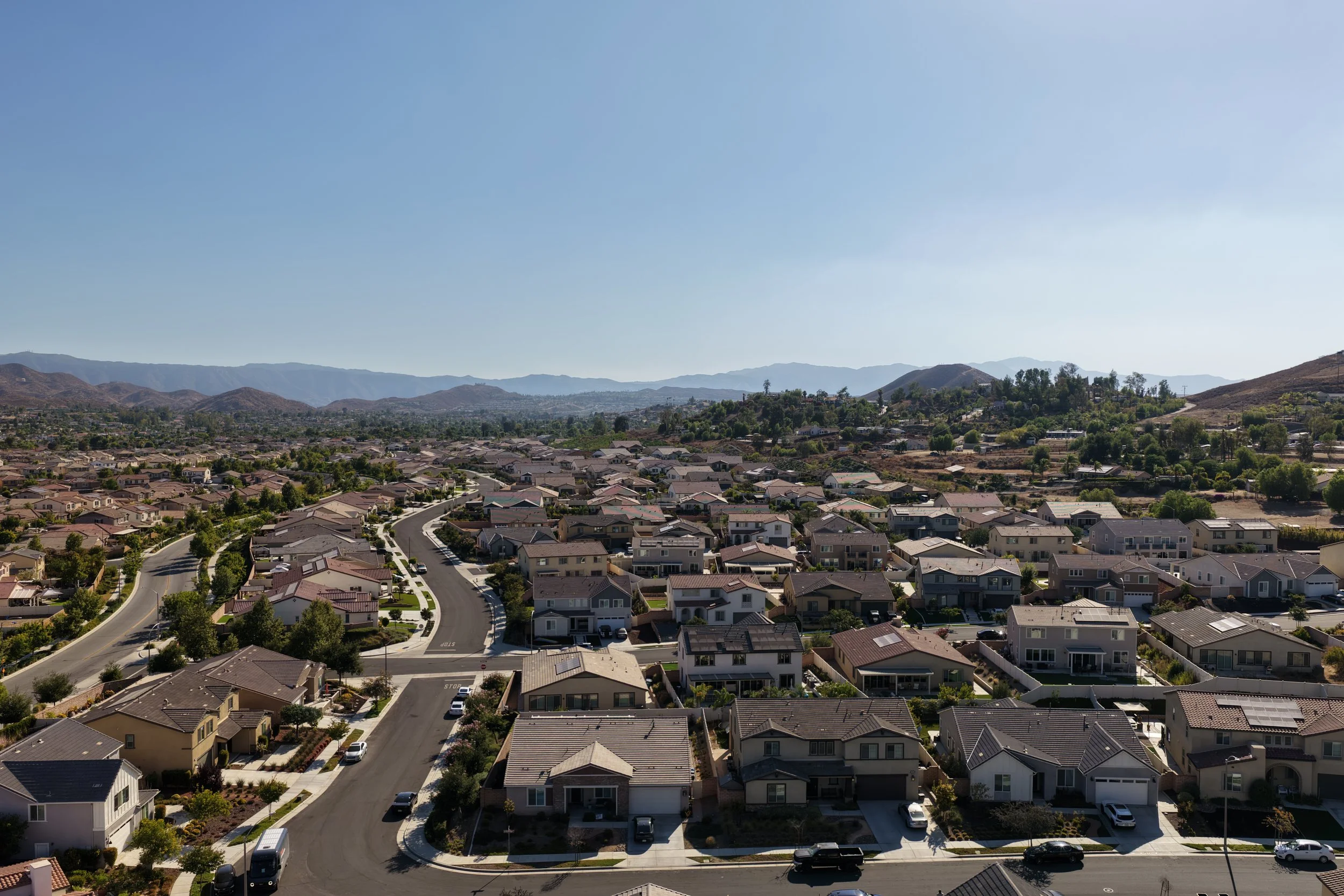 Aerial view of a suburban neighborhood with houses, streets, and greenery, set against a backdrop of mountains and a clear blue sky.
