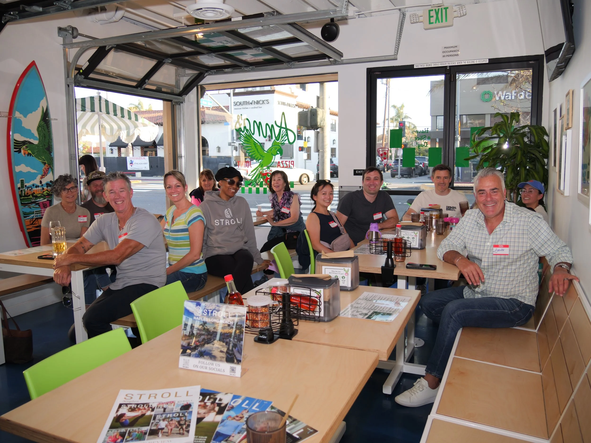 Group of people sitting around tables inside a restaurant, smiling and enjoying their time, with menus and drinks on the tables, and large windows showing the city outside.