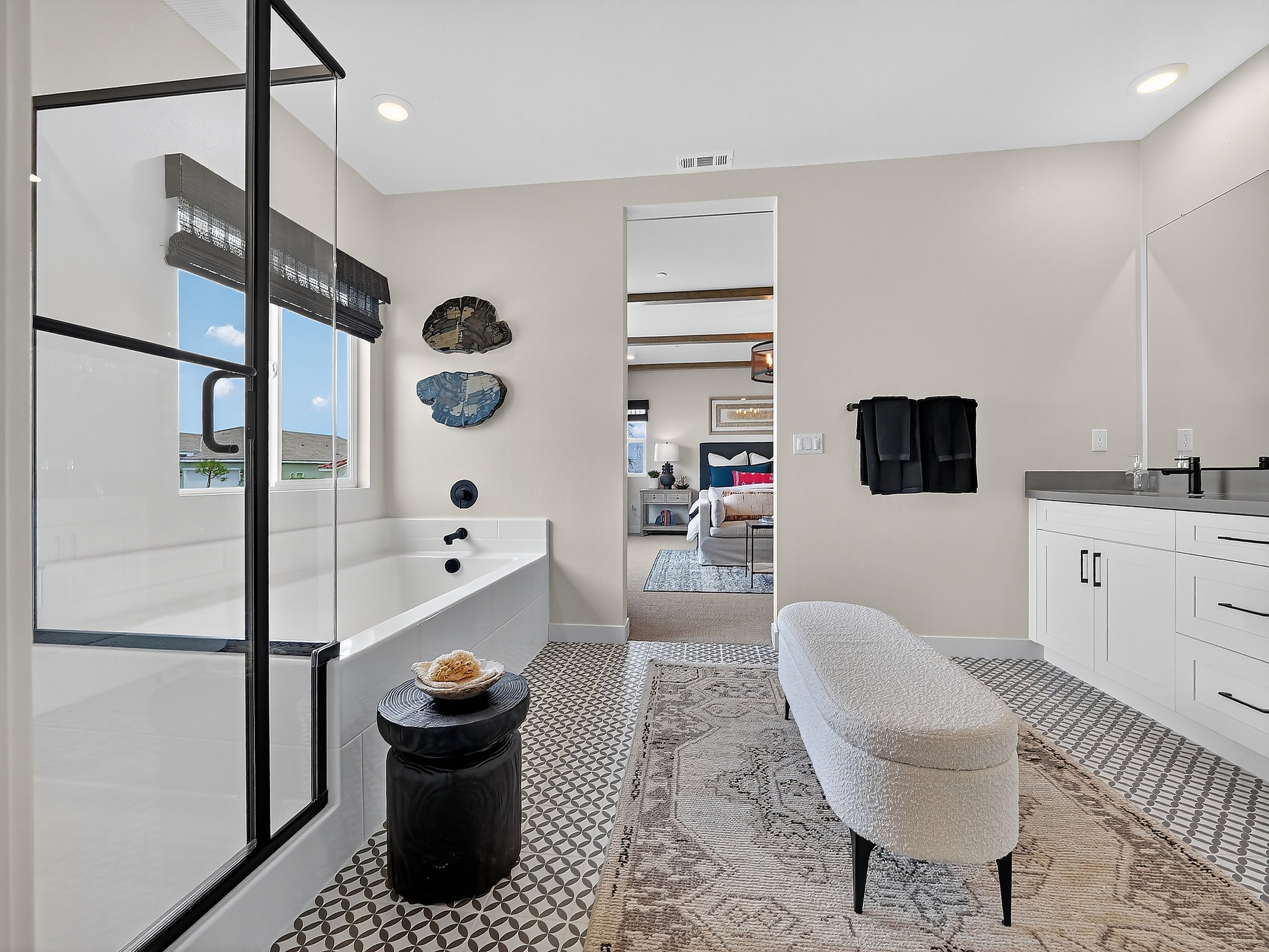 A modern bathroom with a glass shower enclosure, a bathtub, and wall art. The bathroom has a neutral color palette with white walls, patterned floor tiles, and black accents. There is a view into a bedroom with a bed, bedside tables, and lamps.