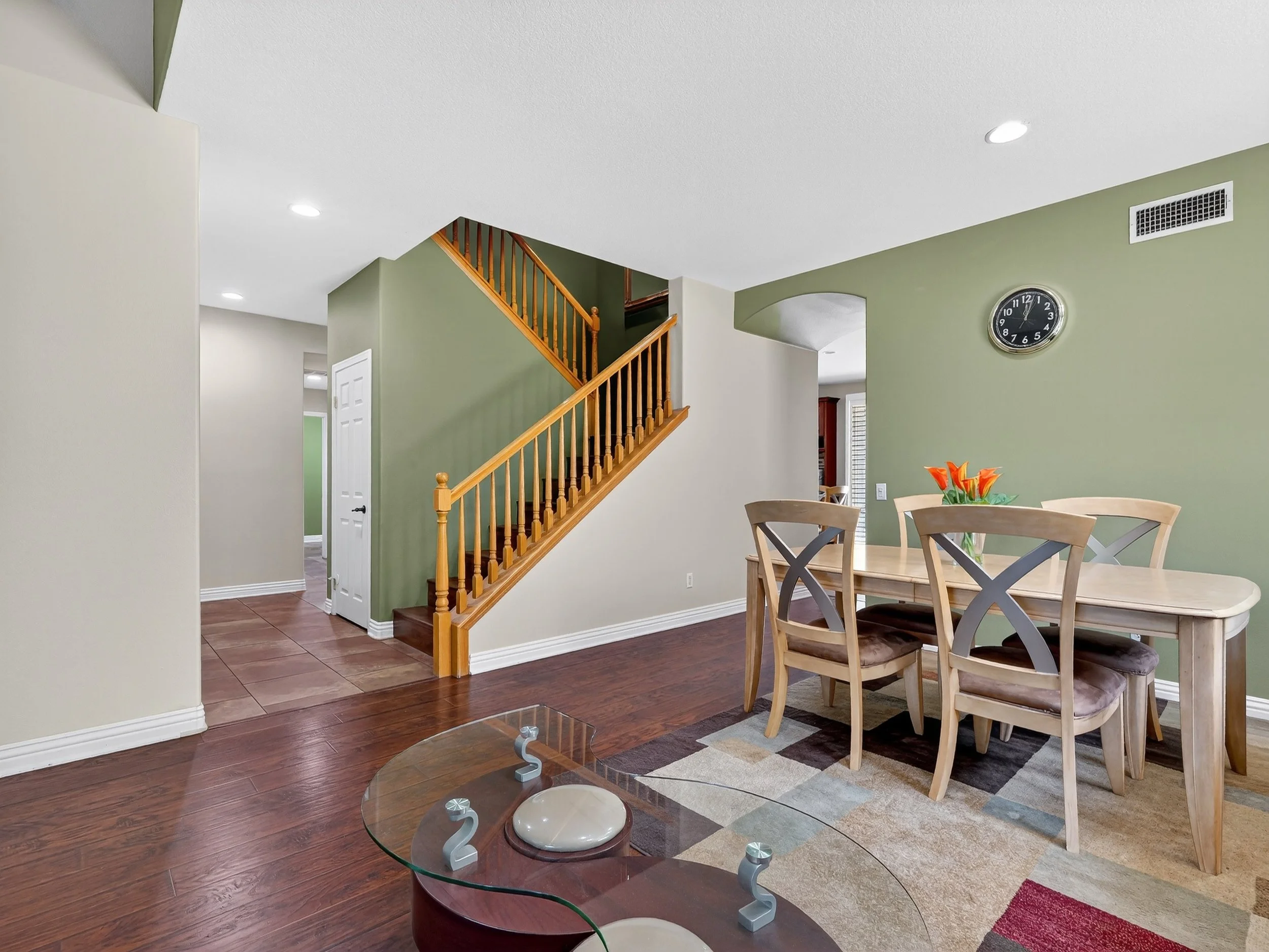 Interior view of a living room with green walls, wooden staircase, and dining table with chairs. A small glass coffee table is in the foreground. A wall clock and ventilation grate are visible on the wall.
