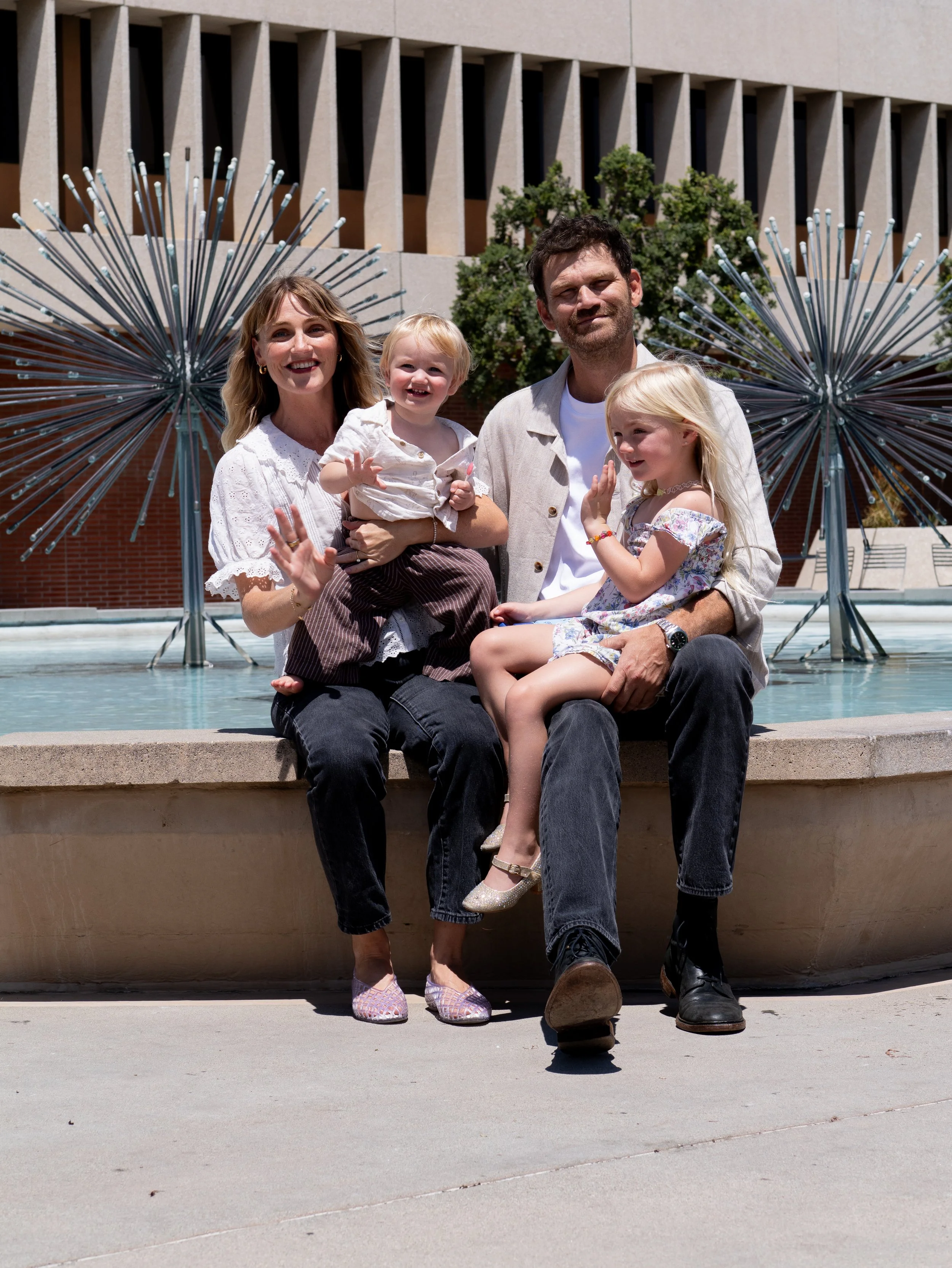 A family of five, including two men, two women, and a young girl, sitting on the edge of a fountain outdoors with modern architecture and large decorative metal art in the background.