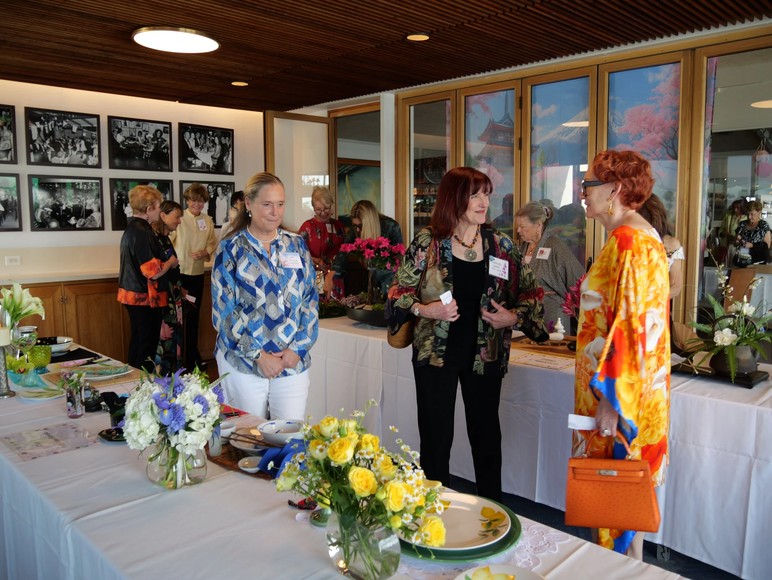 Group of women at an indoor craft or flower arranging event with tables decorated with yellow and purple flowers, antique black and white photographs on the wall, and large windows with scenic views.