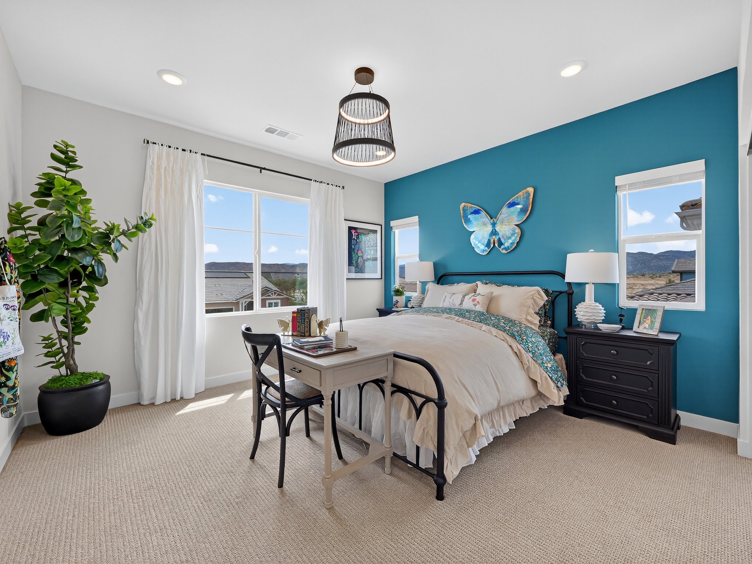 A bedroom with a blue accent wall, large windows with white curtains, a black bed frame, a cream-colored bedspread with floral and patterned pillows, black nightstands with white lamps, and a framed butterfly wall art. There is a tall potted plant ne