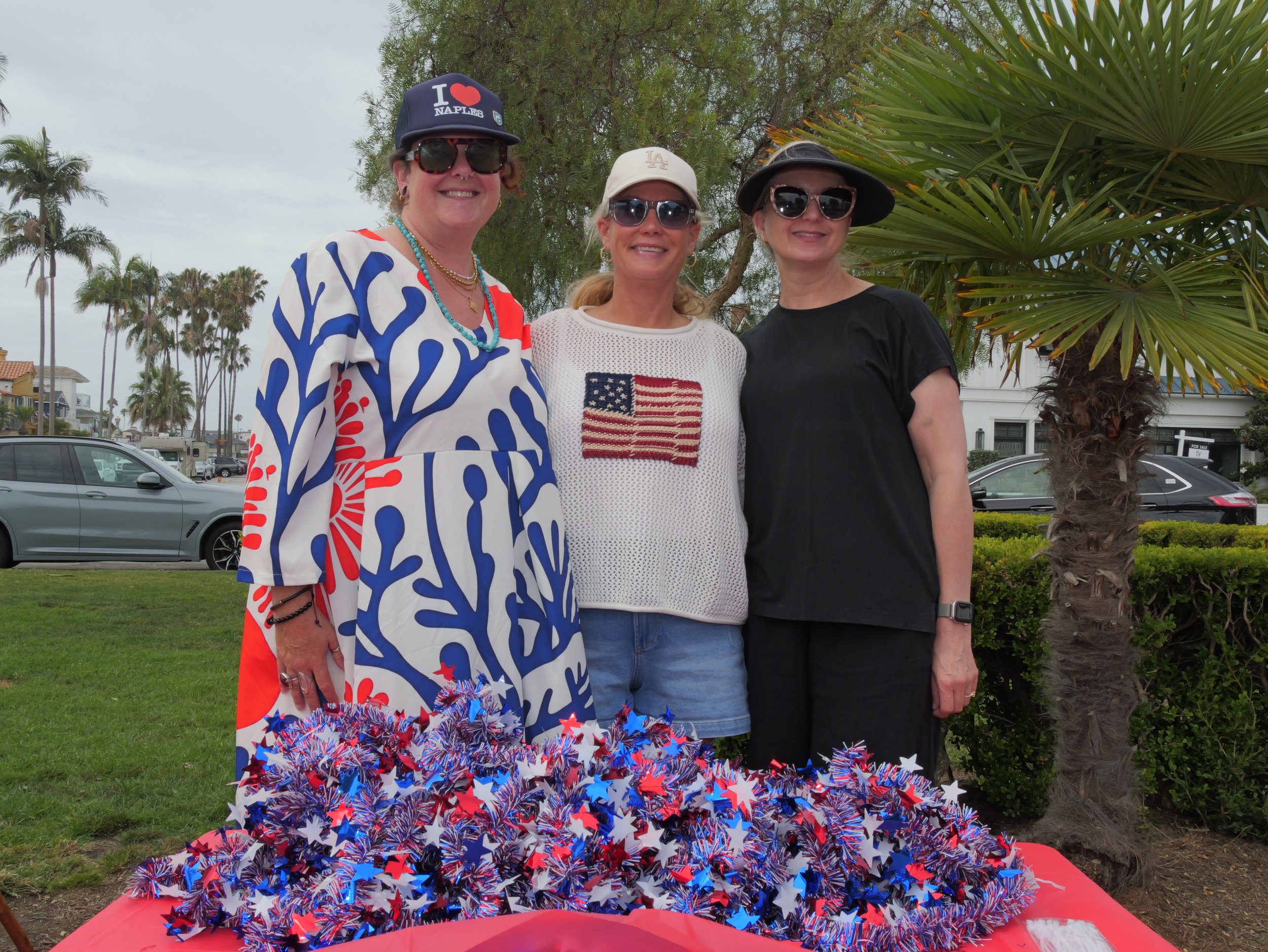 Three women standing outdoors near a palm tree, celebrating a patriotic event with red, white, and blue star-shaped decorations on the ground. All three women are wearing sunglasses and smiling, dressed in casual clothing with patriotic themes.