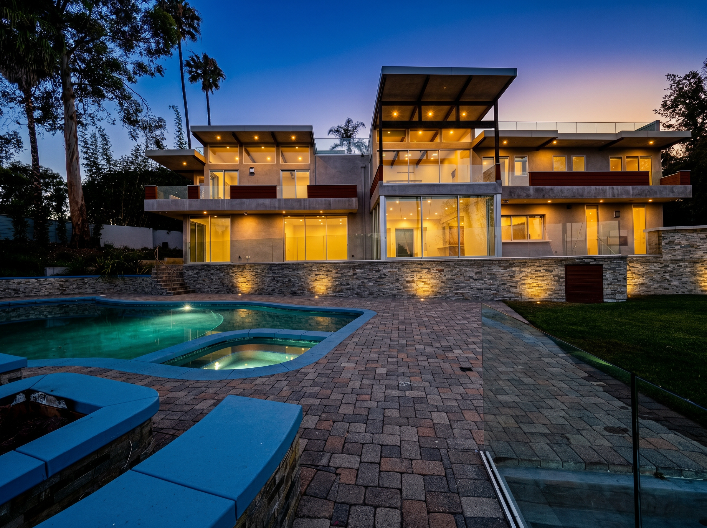 Modern multi-story house with large glass windows, outdoor lighting, and a swimming pool in the backyard during twilight.