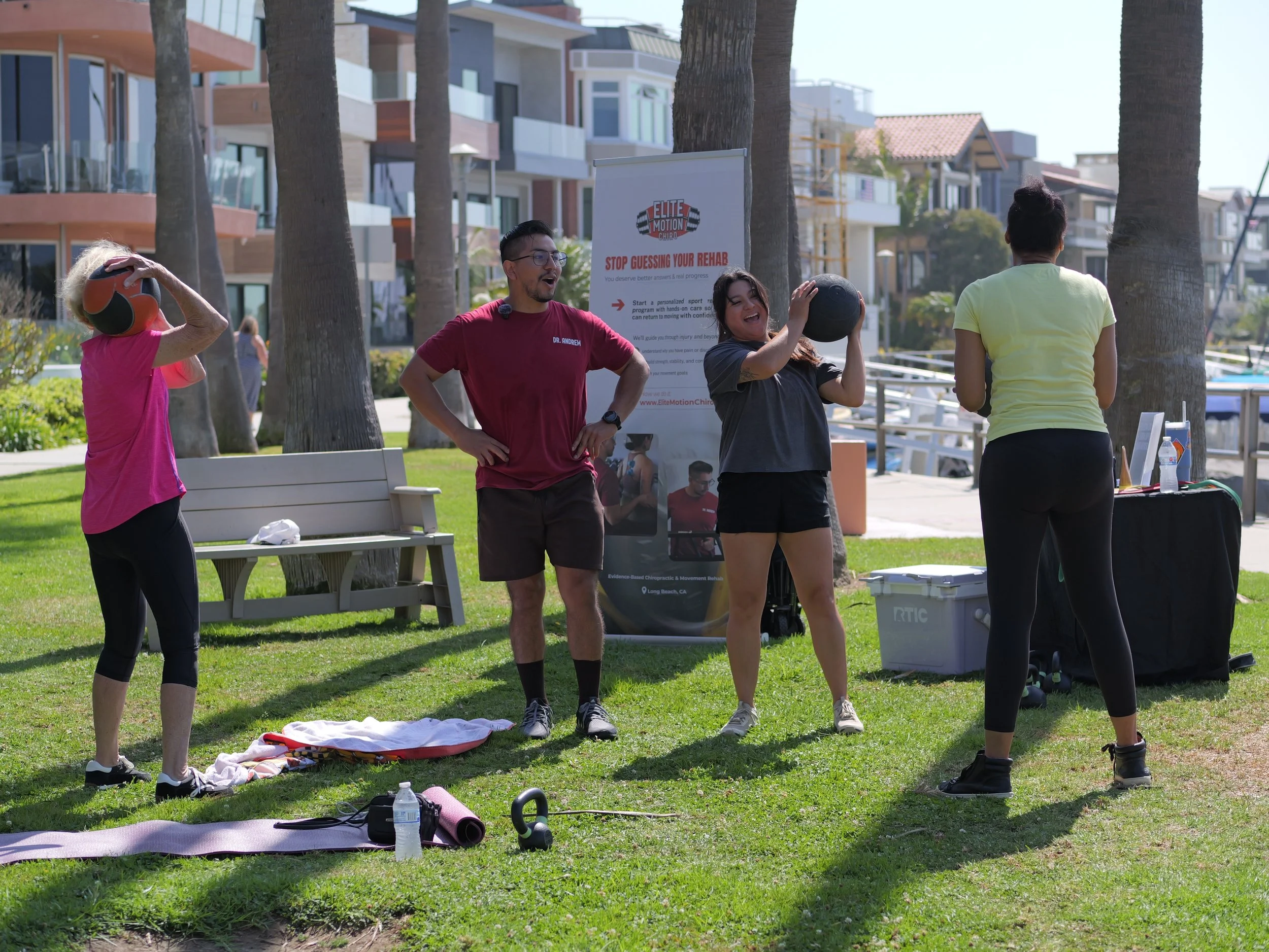 Four individuals engaging in outdoor fitness training or physical therapy session in a park, with one person holding a medicine ball, in front of a sign for Elite Motion Chiropractic, with residential buildings and palm trees in the background.