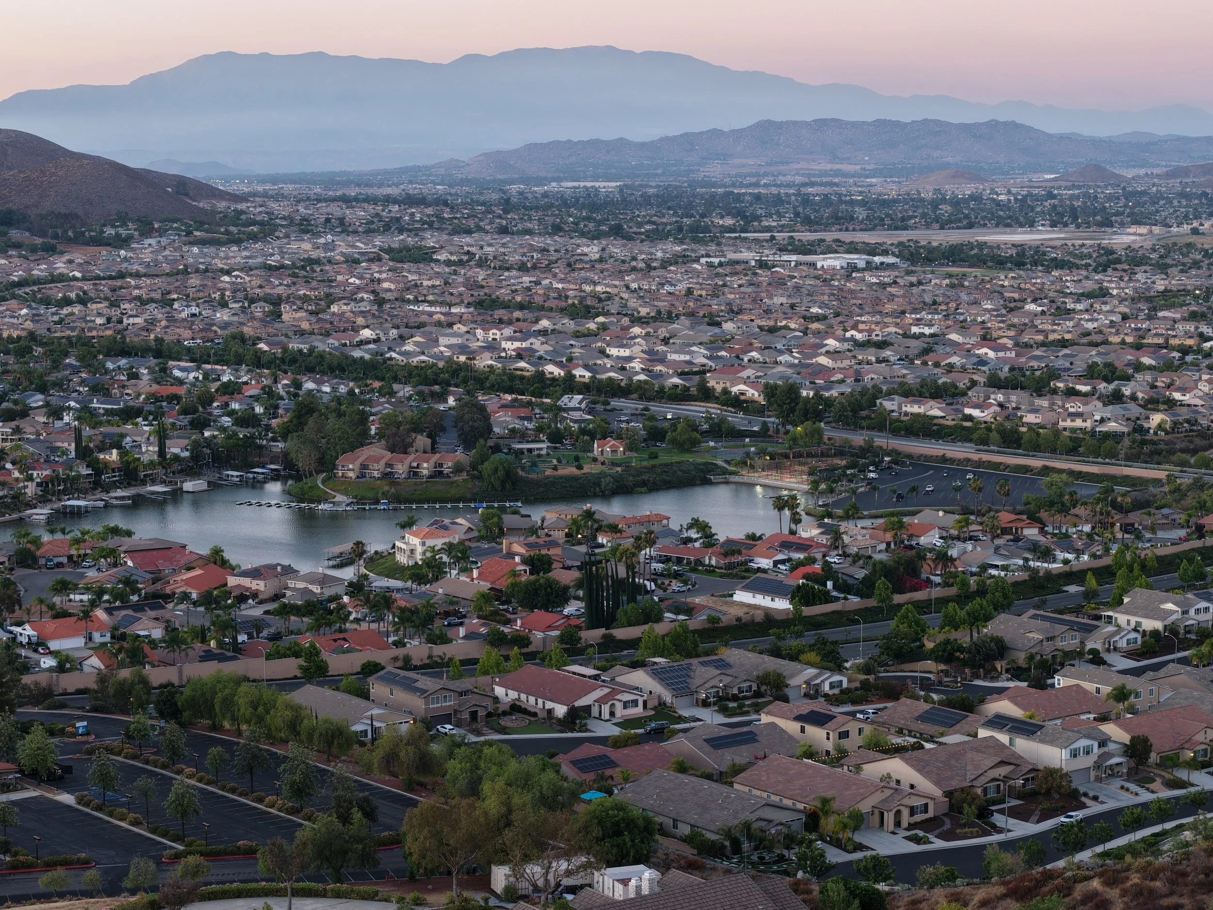 Aerial view of a suburban neighborhood with numerous houses, trees, a small lake, and mountains in the background during twilight.