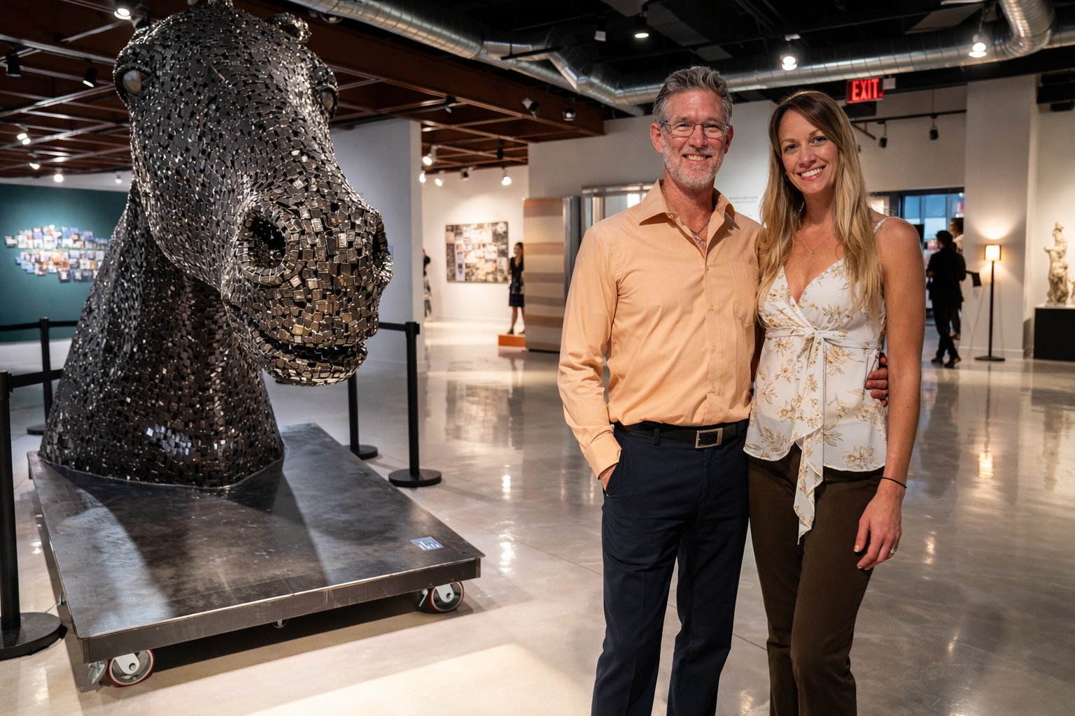 A man and a woman standing in an art gallery, smiling at the camera, with a large silver sculpture of a horse's head made from small, reflective pieces beside them.
