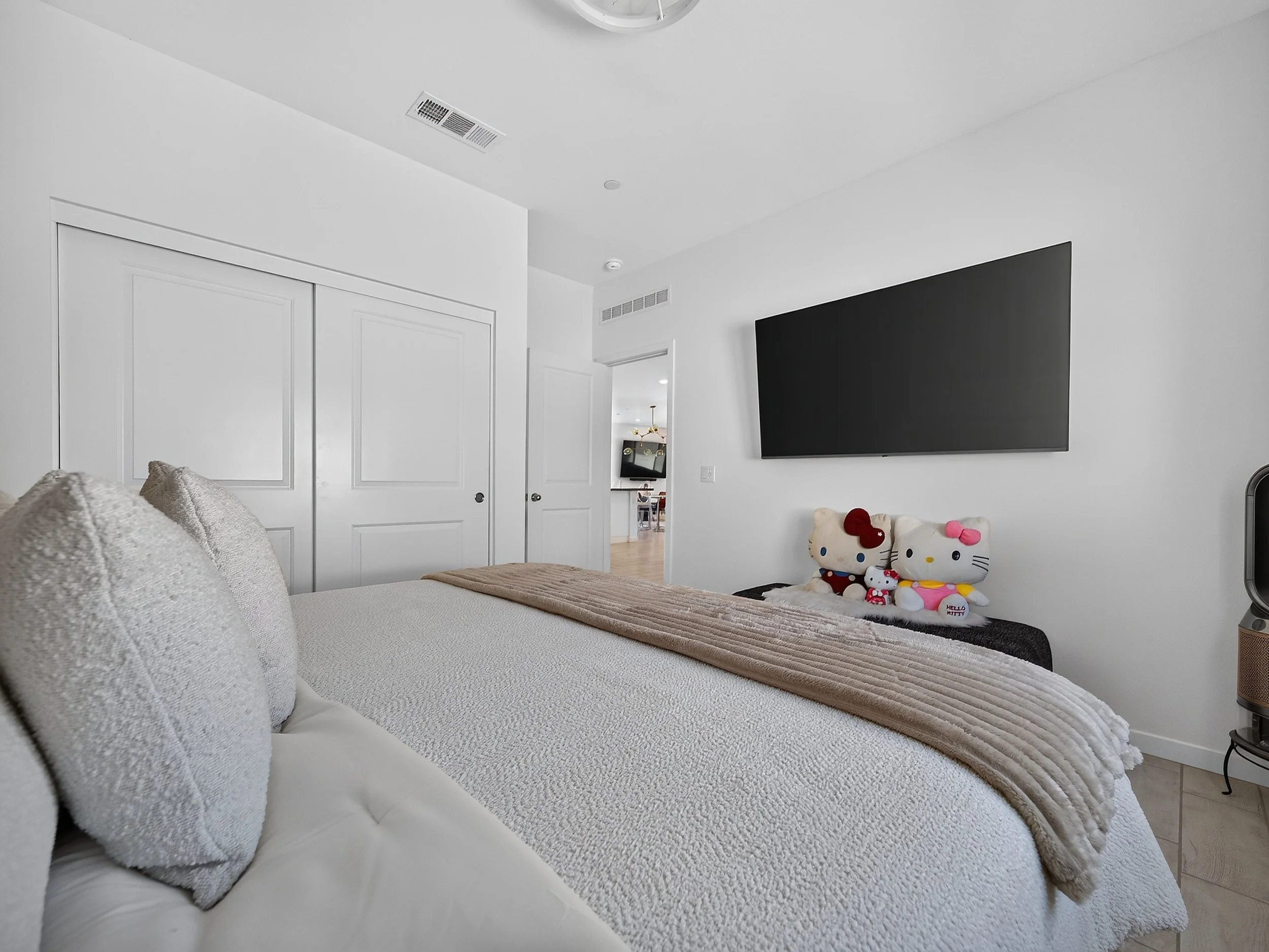 A living room with white walls and a white ceiling, a large TV mounted on the wall, plush white cushions on a beige sofa, and a collection of Hello Kitty plush toys on a side table.