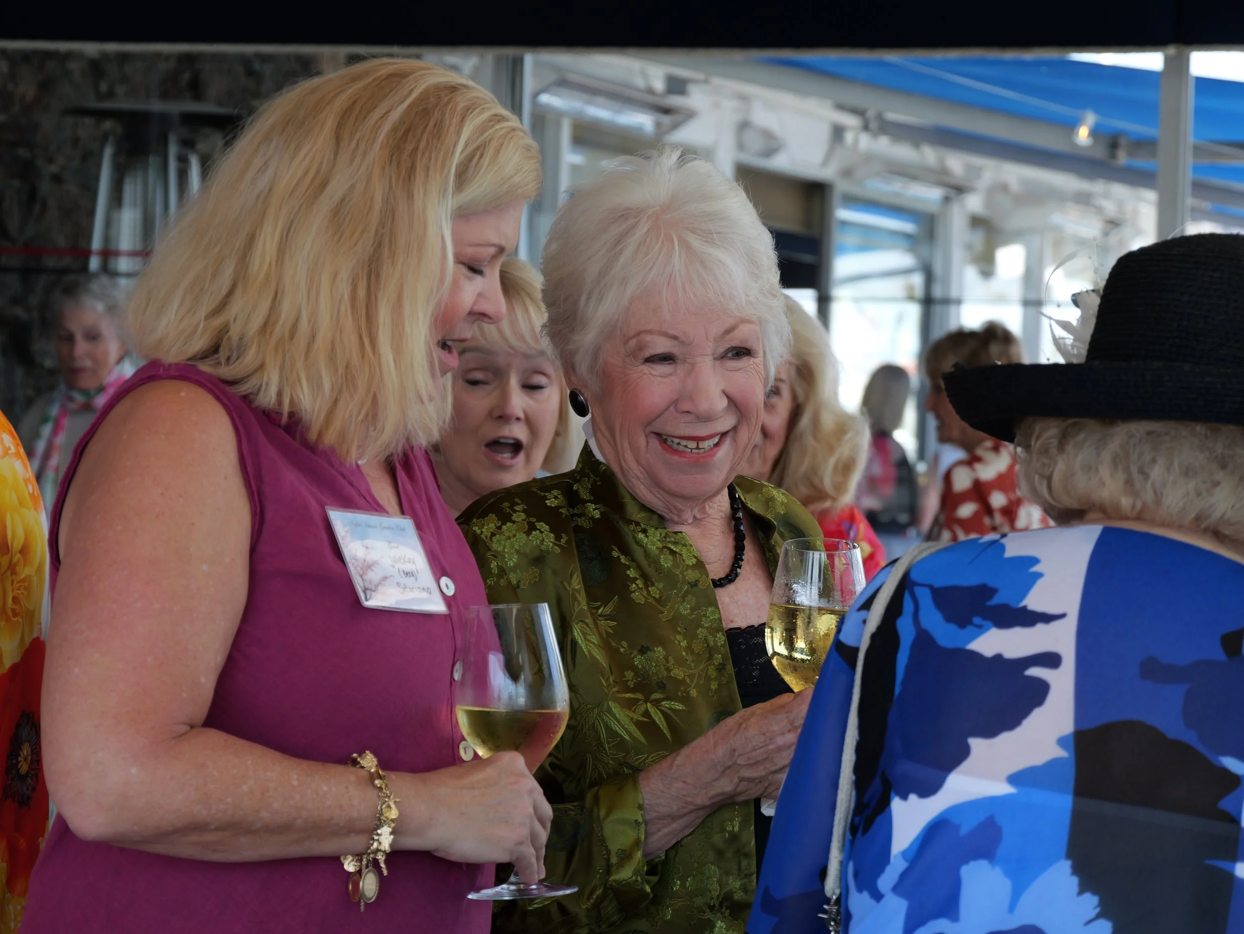A group of women gathered at a social event, holding glasses of white wine, engaged in conversation, with one woman smiling widely.