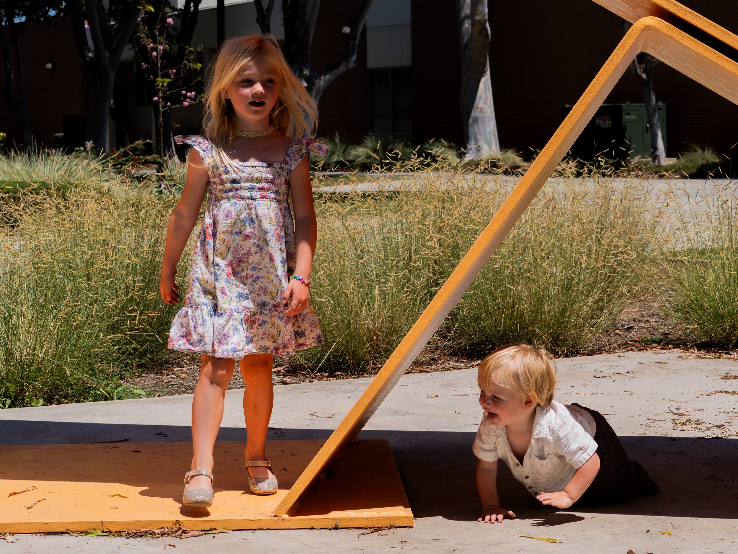 A young girl with blonde hair wearing a floral dress and jewelry standing outside near a wooden structure while a young boy with blonde hair in a white shirt and dark striped pants is crawling underneath it.