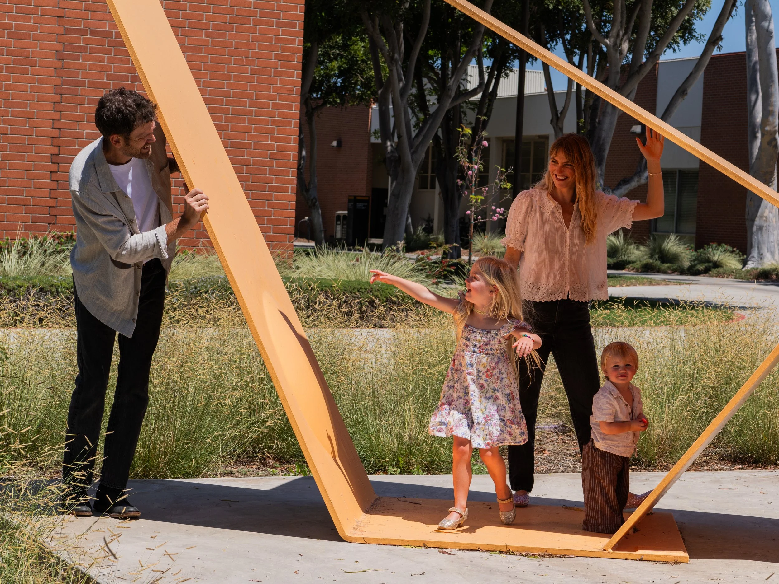 A man and a woman with two young girls playing on a large, orange, abstract wooden sculpture outdoors. The man is smiling and leaning on the sculpture, the woman is smiling and waving, one girl is pointing, and the other girl is standing near the scu