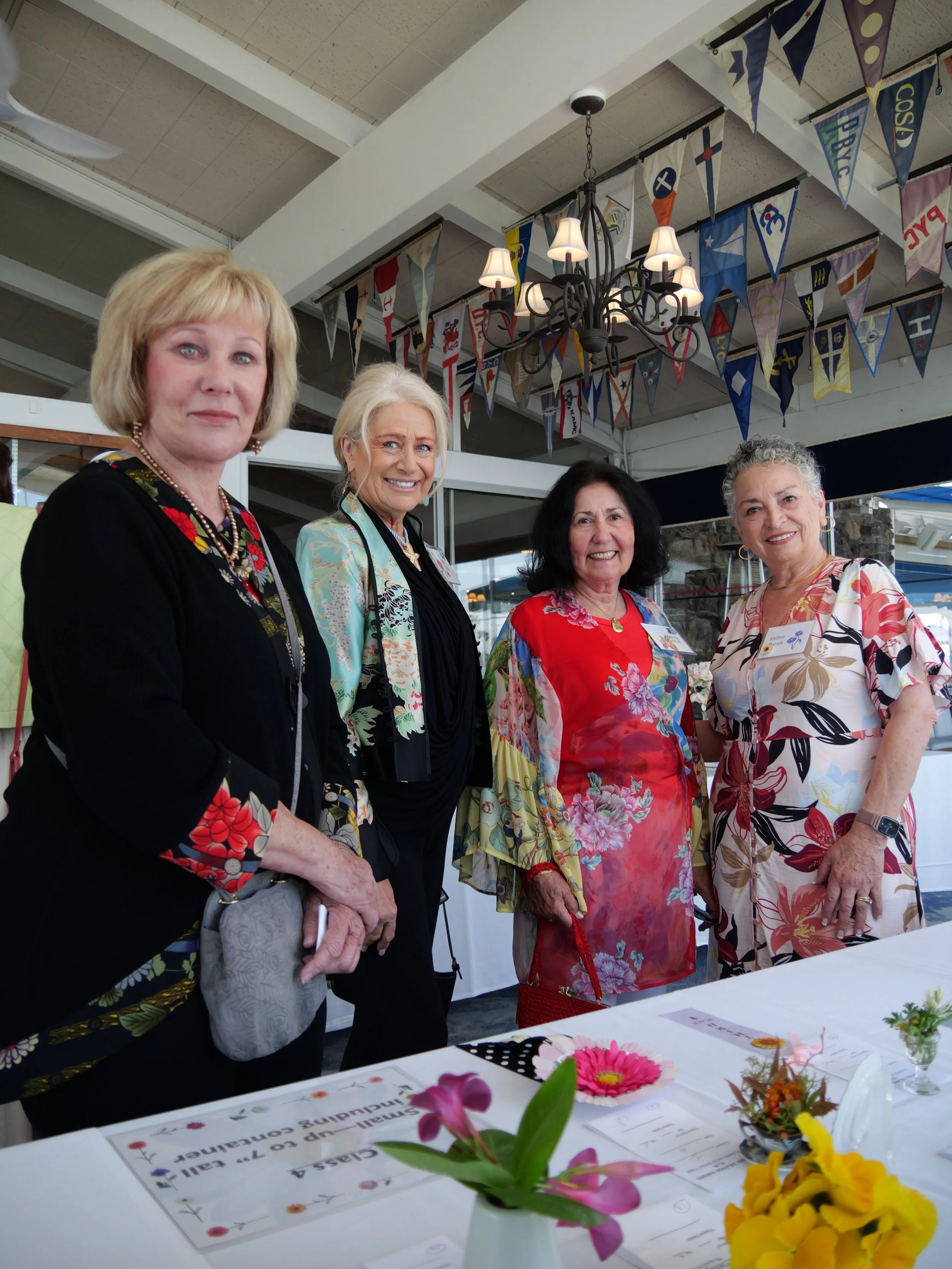 Four women standing behind a table decorated with flowers, with banners hanging from the ceiling in the background.