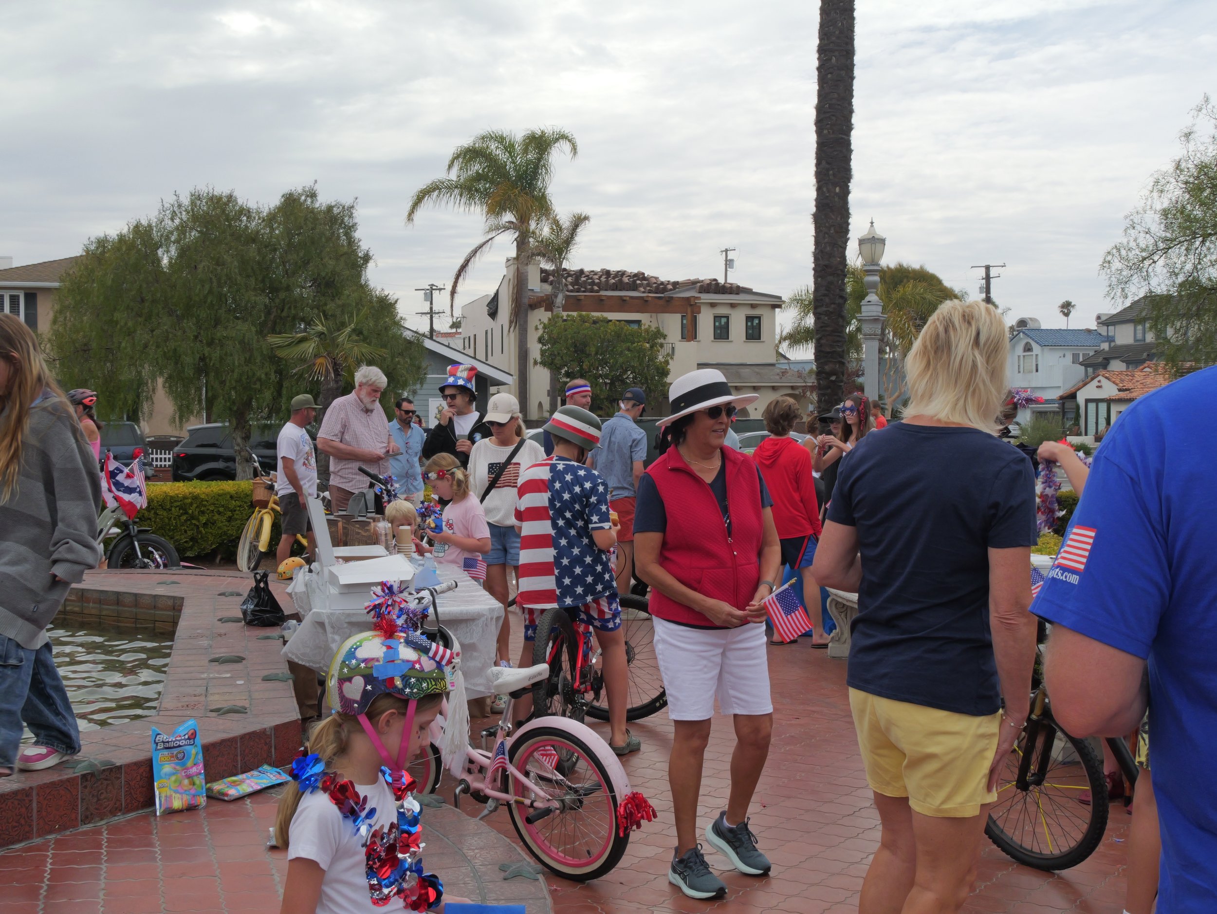 People gathered near water during a patriotic celebration, wearing red, white, and blue attire with American flags and decorations, with houses, trees, and cloudy sky in the background.