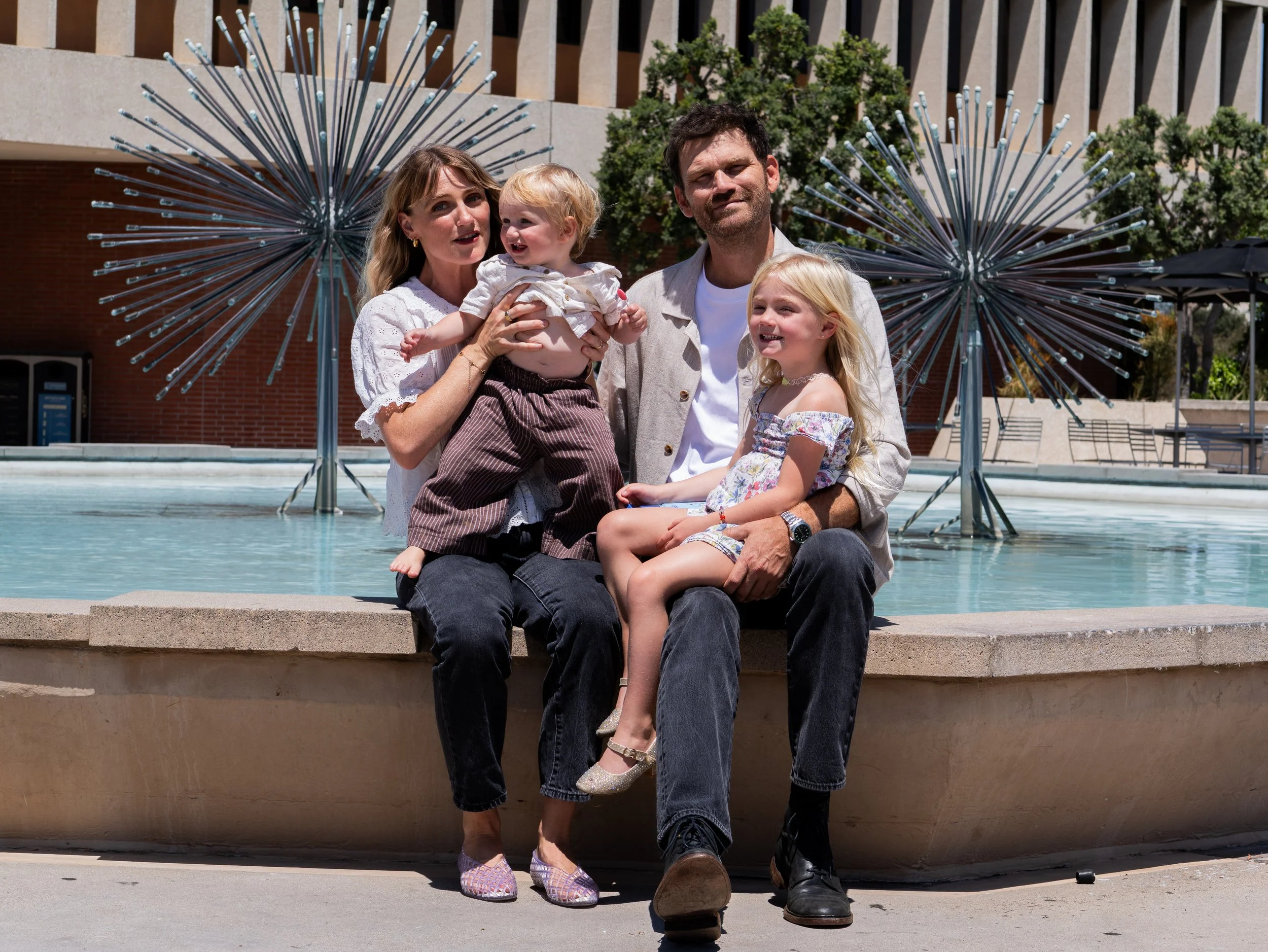 A family of four, including a woman, a man, and two young girls, sitting together on a fountain edge outside in a sunny urban area with modern building and greenery in the background.