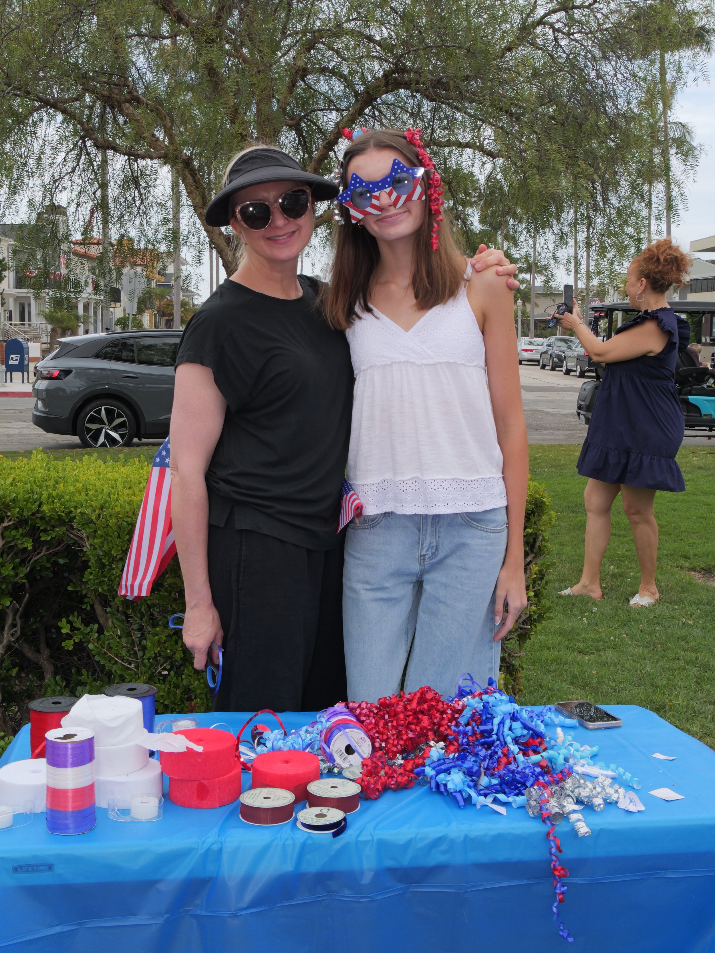 Two women standing together outdoors with a table of patriotic decorations and accessories in front. They are smiling, wearing festive sunglasses, and celebrating. One woman has a black hat and black shirt; the other has novelty sunglasses with an Am