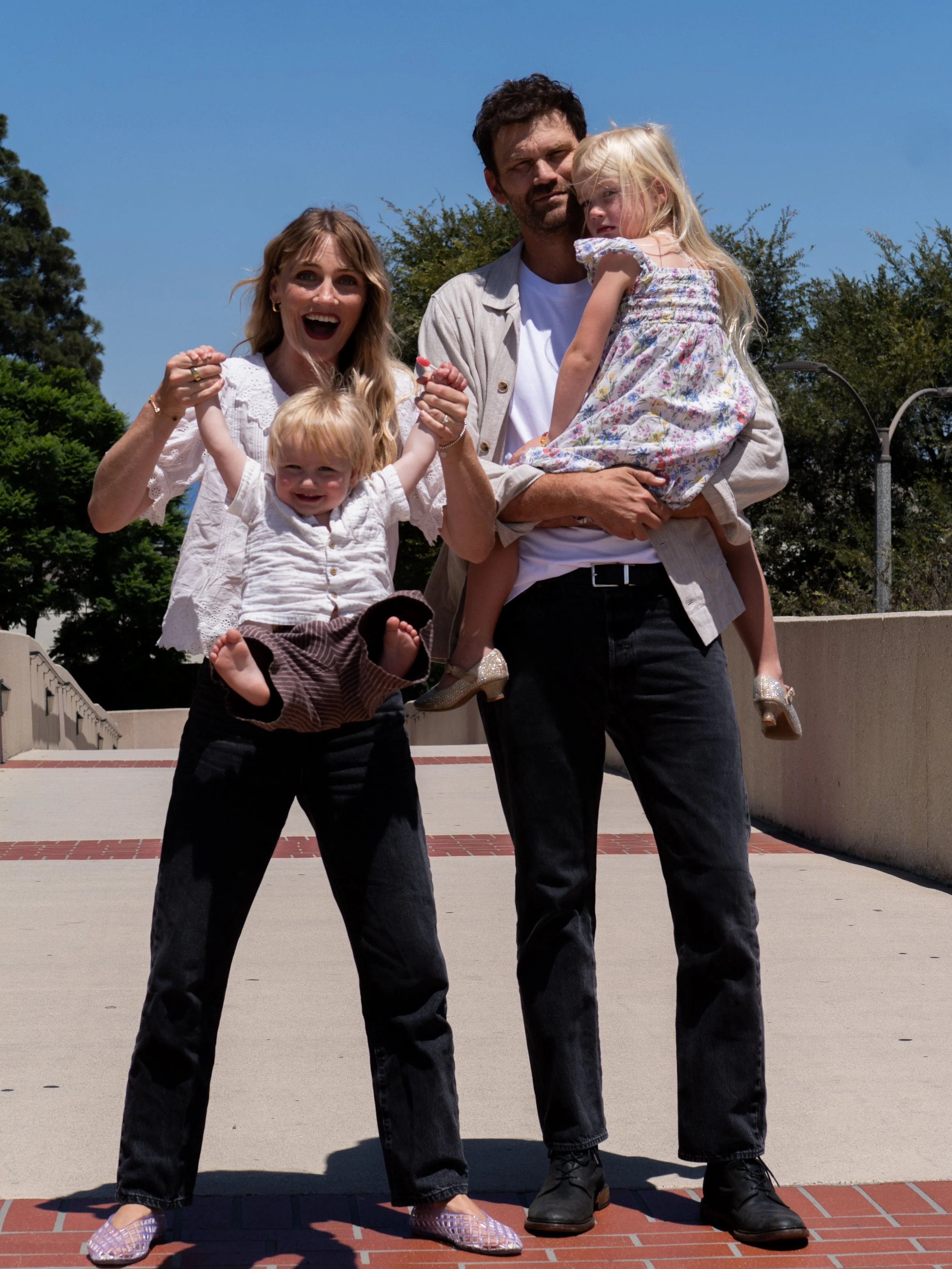 A family of four outdoors on a sunny day, with a woman, a man, and two young girls. The woman is smiling and holding one girl with blonde hair and a white shirt. The man is holding the other girl, who is dressed in a floral dress. The group is standi
