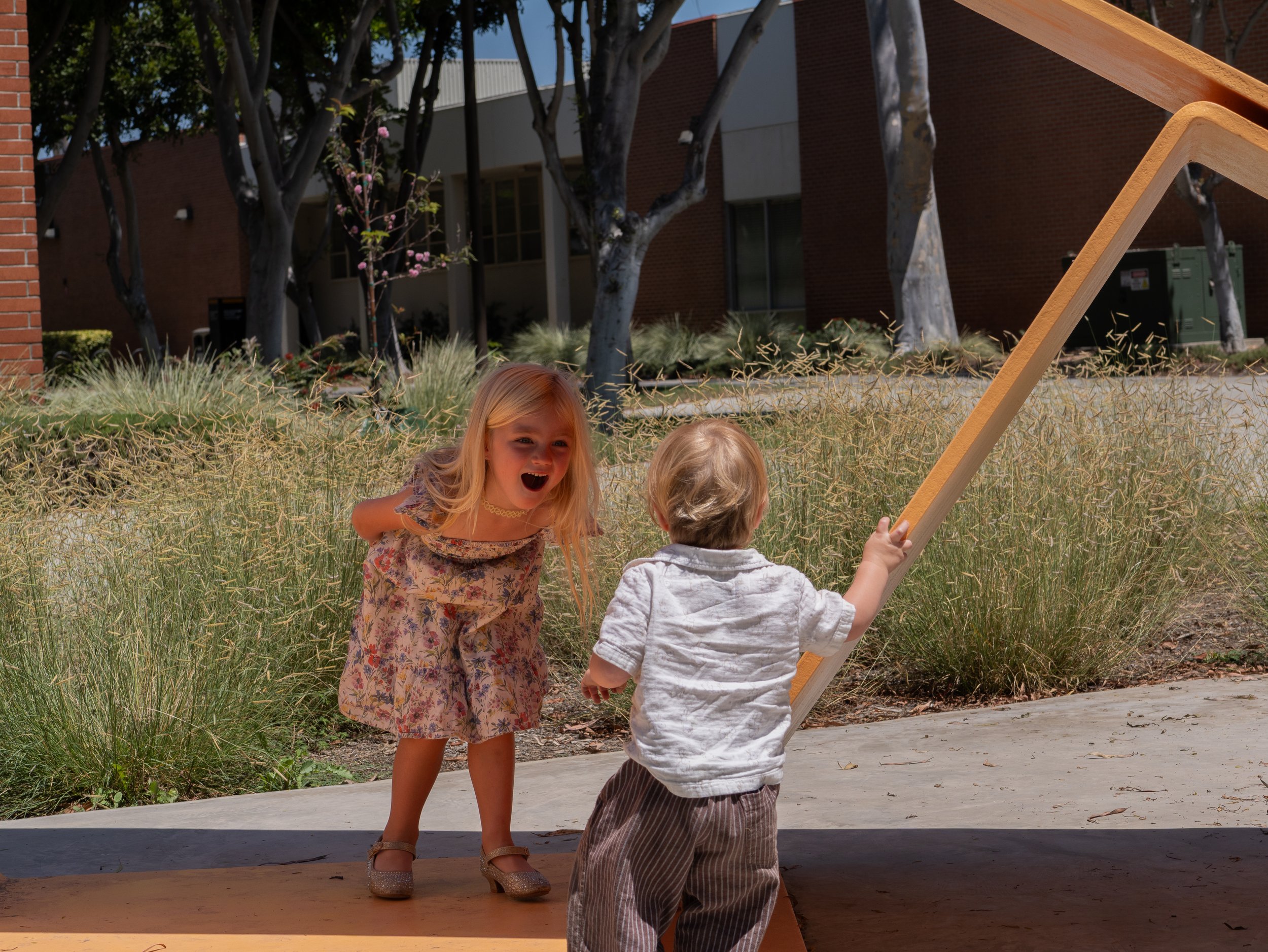 Two young children, a girl with long blonde hair and a boy with short light hair, playing outdoors near a wooden structure, with trees and a building in the background.