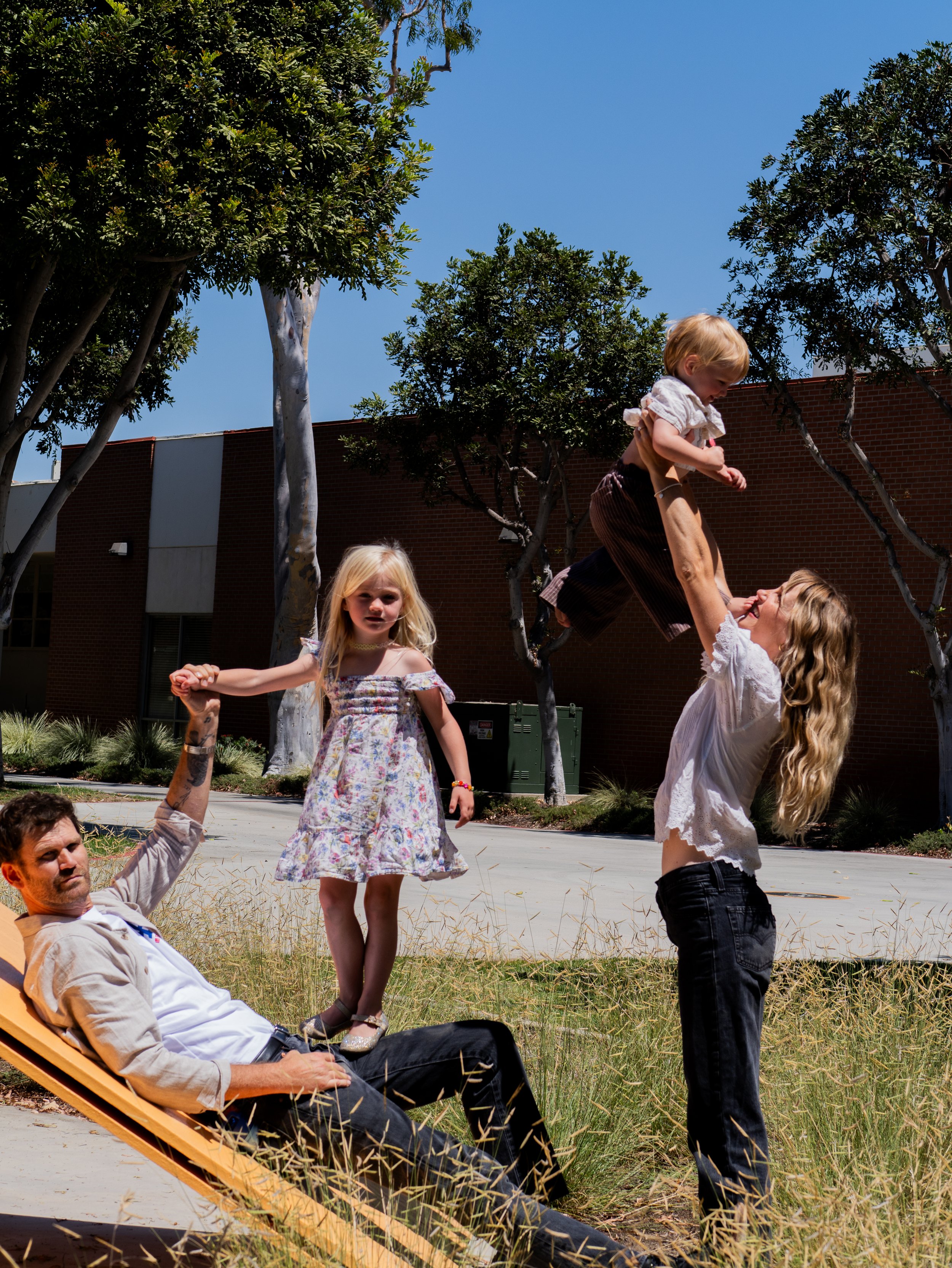 A group of people, including children, enjoying an outdoor activity on a sunny day, with some holding hands and others interacting, surrounded by trees and grass.