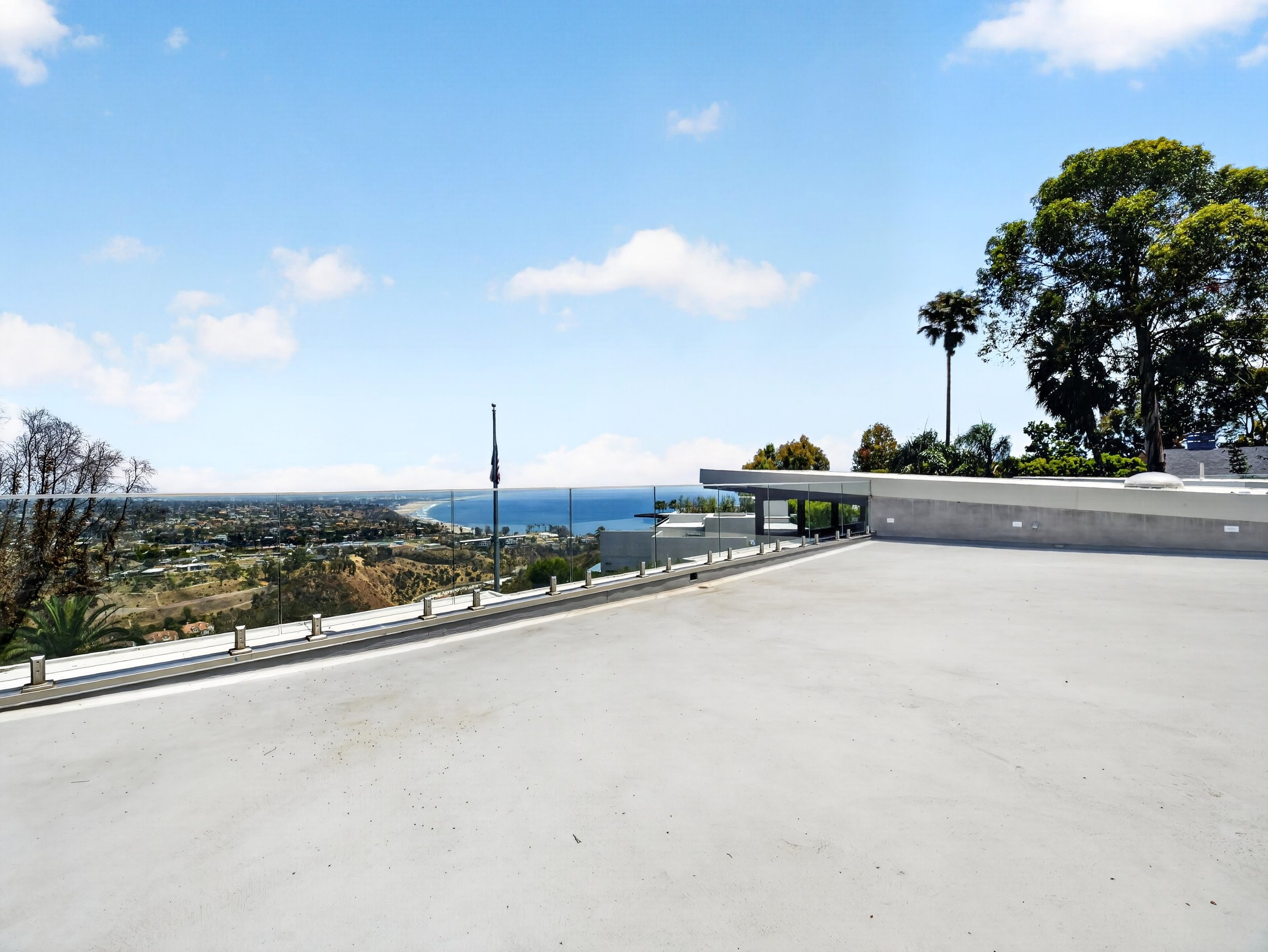 Empty rooftop balcony with a glass railing, overlooking a cityscape, with palm trees and a blue sky with clouds.