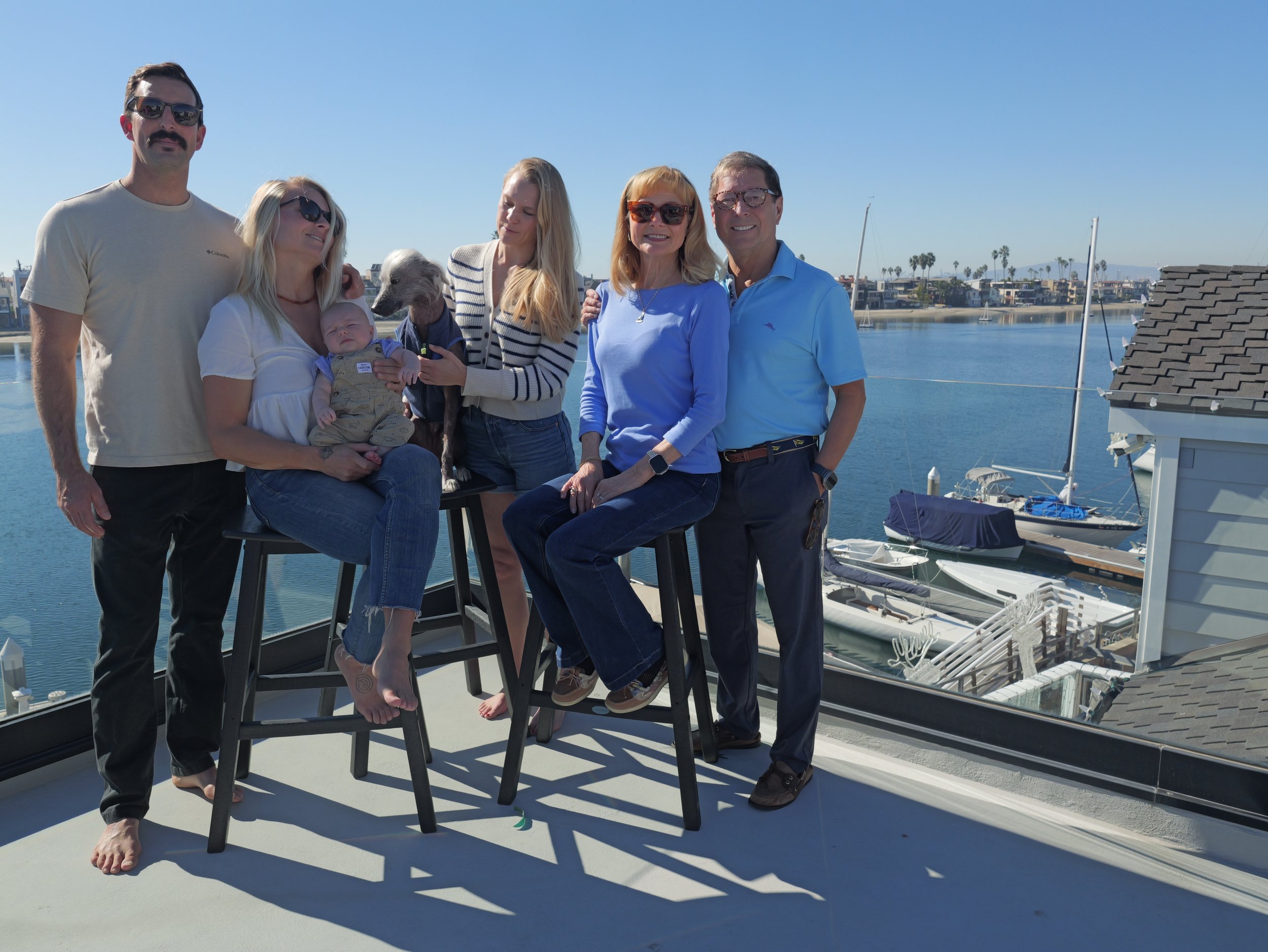 A group of seven people and a dog on a balcony overlooking a marina with sailboats and palm trees.