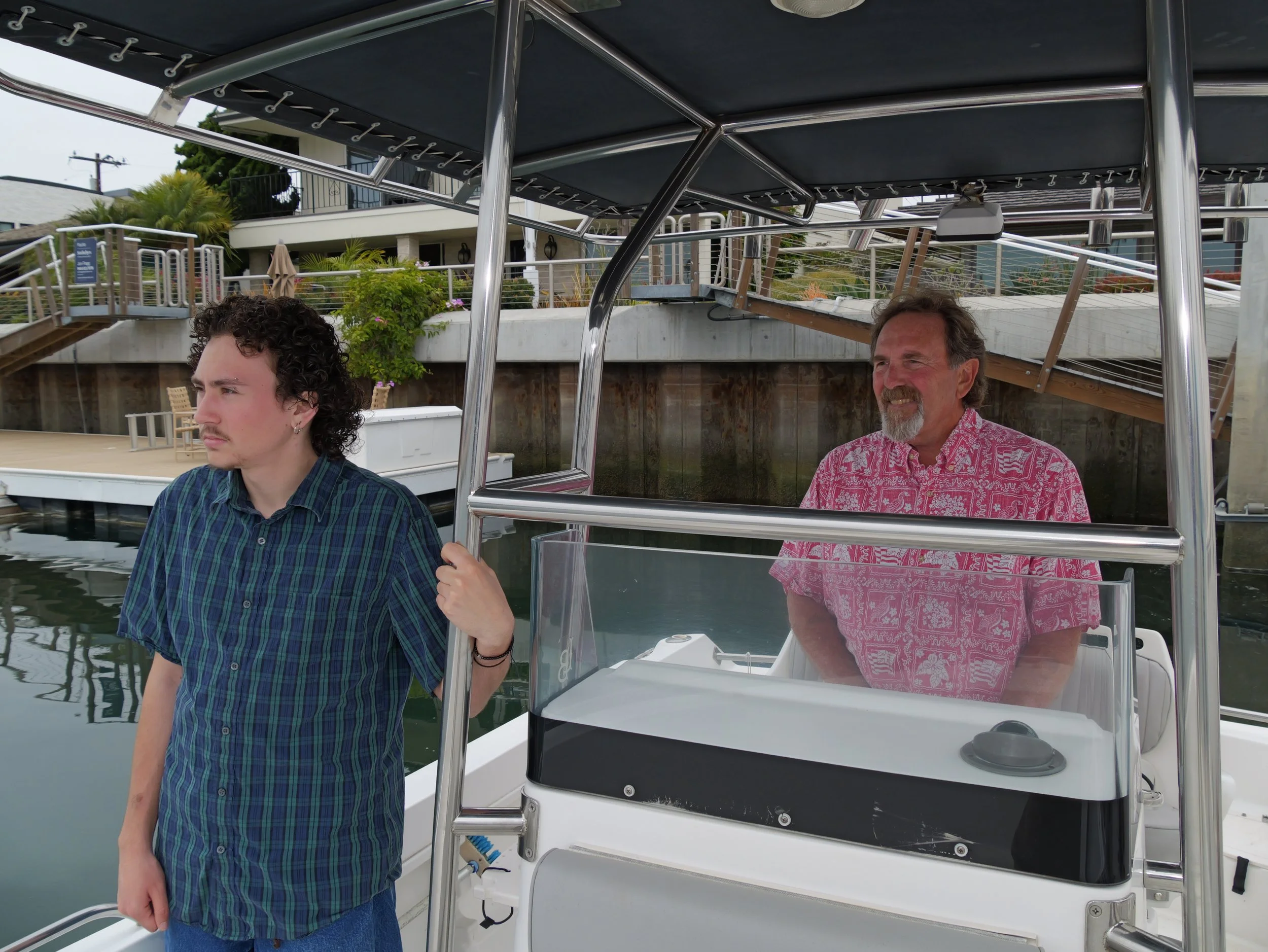 Two men on a boat docked near a residential area with greenery and modern buildings. The younger man in front has curly hair and is wearing a plaid shirt, while the older man in back has a beard and is wearing a pink patterned shirt.