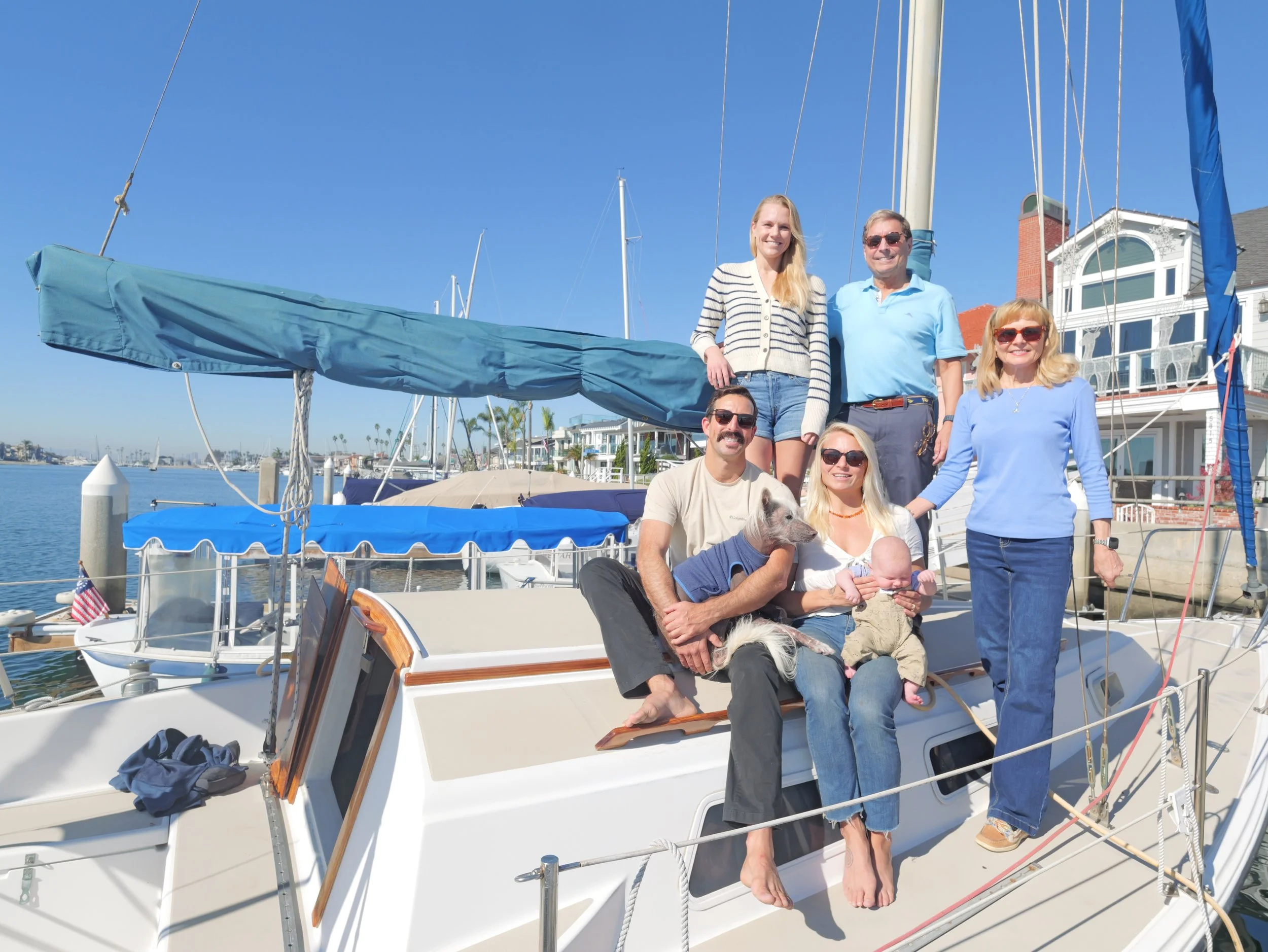 Group of six people, four women and two men, on a boat docked at a marina, smiling and posing for the photo with clear blue skies in the background.