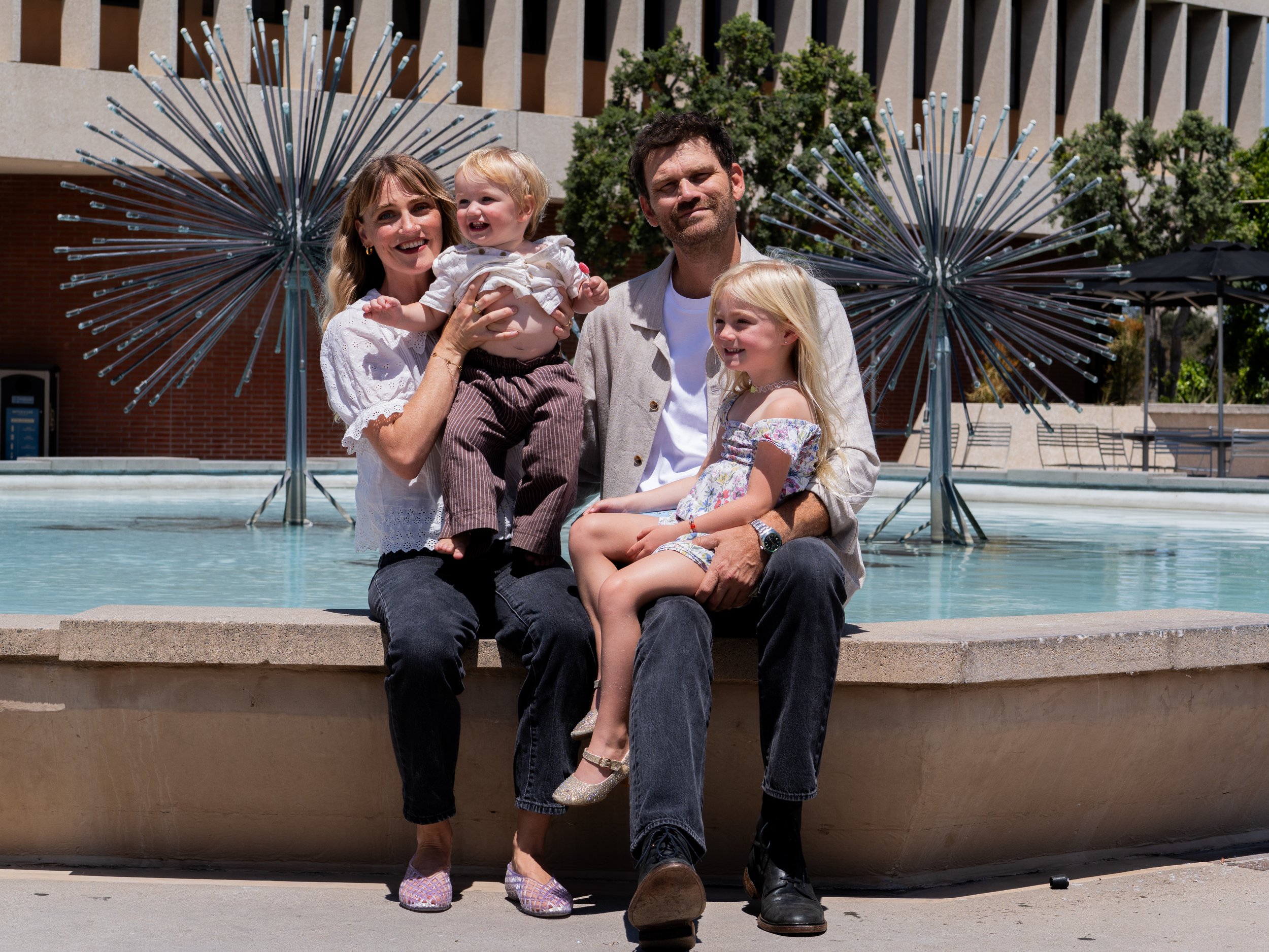 Family of four sitting by water fountain outdoors, with modern building and trees in background. Woman holding a young child, man sitting with girl, all smiling.