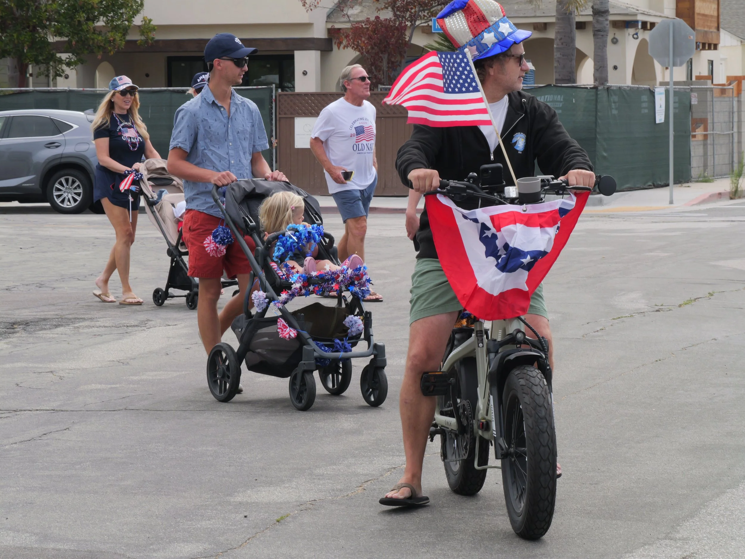 Man riding an electric bike decorated with American flags, leading a parade. Behind him, a woman, a man, and two young children walking, all wearing patriotic clothing and accessories, celebrating a holiday in a neighborhood.
