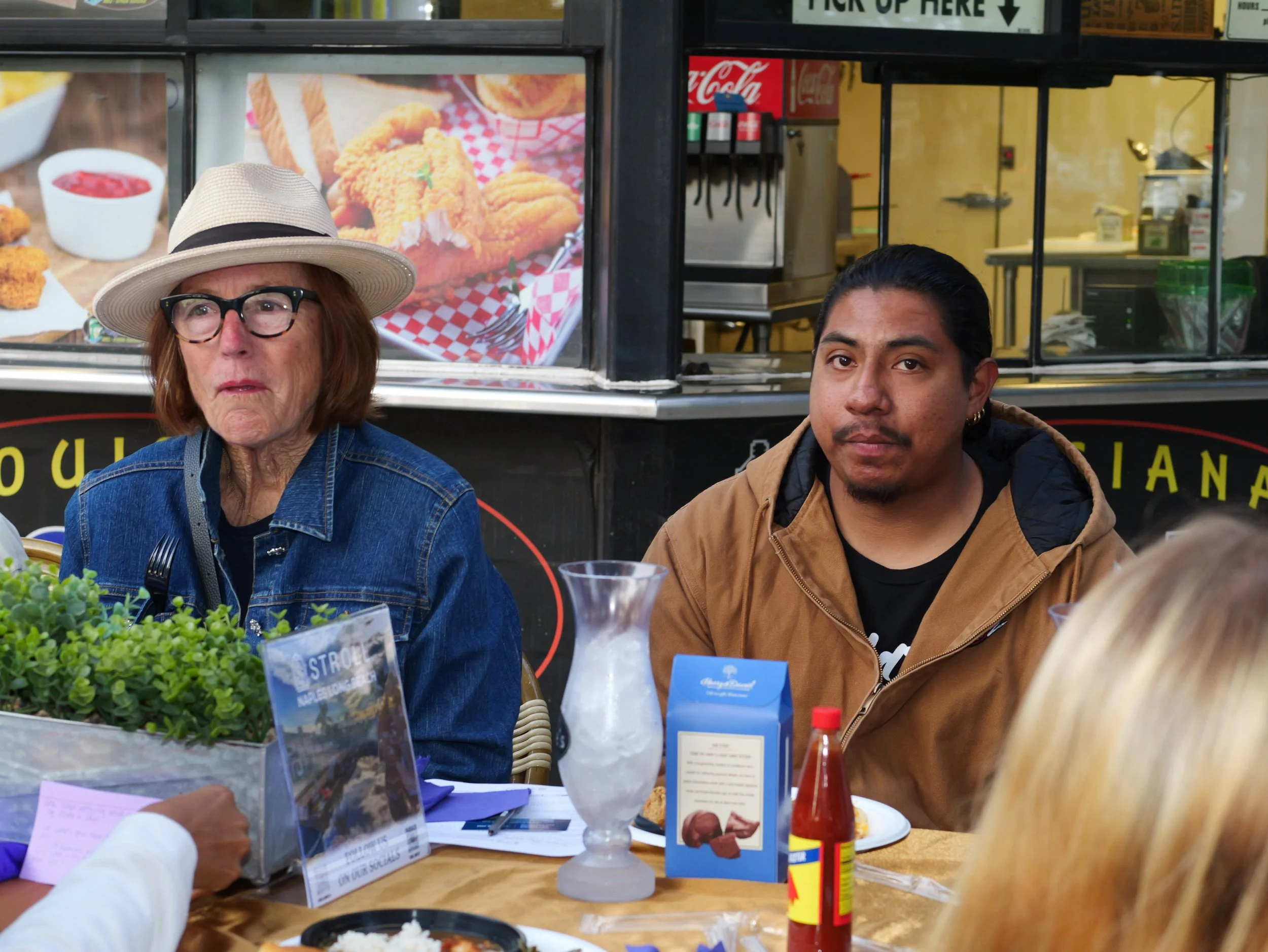 Two people sitting at a food stand table, with a woman's face partially visible on the left and a man with dark hair, light brown skin, and a brown jacket on the right. The background shows a food menu and a food truck with a large image of fried chi