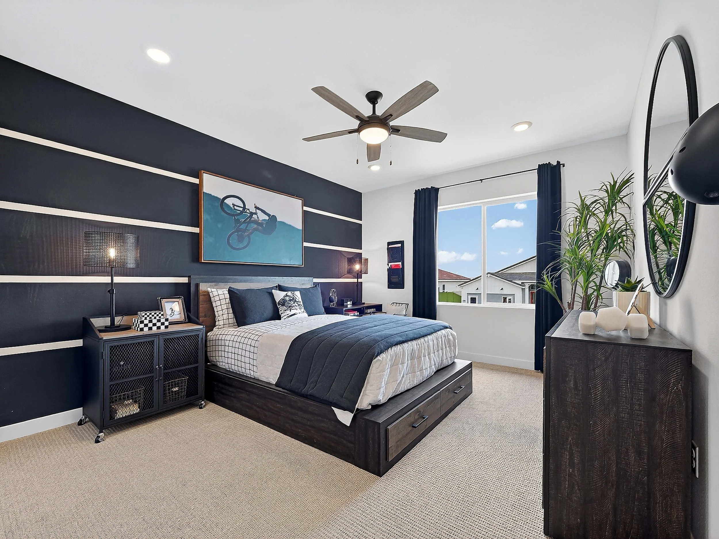 A modern bedroom with a black and white color scheme, featuring a large bed with checkered and solid pillows, a black bedside table with a lamp, a ceiling fan, and a window with blue curtains, complemented by indoor plants and wall art.
