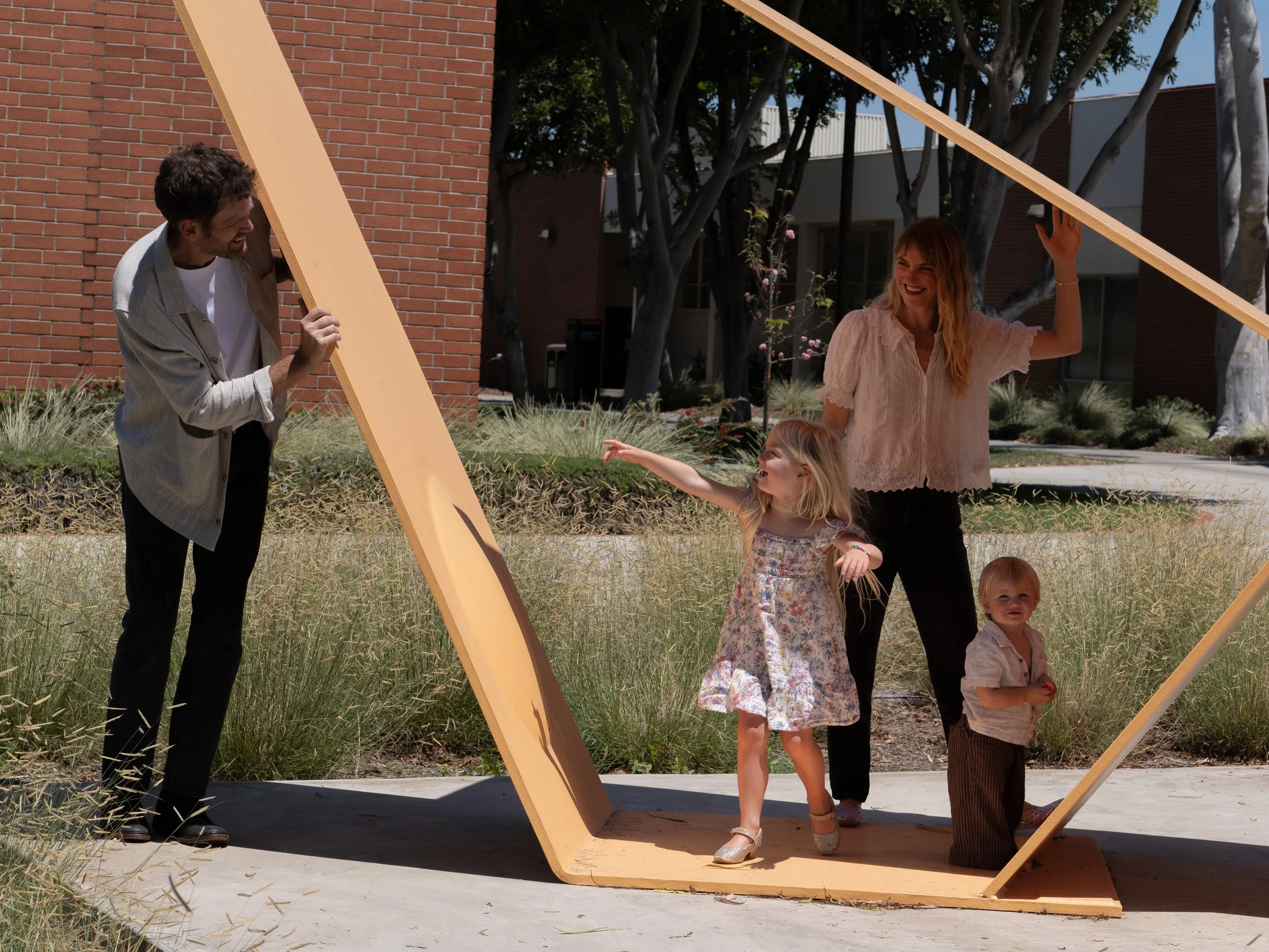 People standing and smiling on a wooden sculpture structure outdoors, with children and trees in the background.