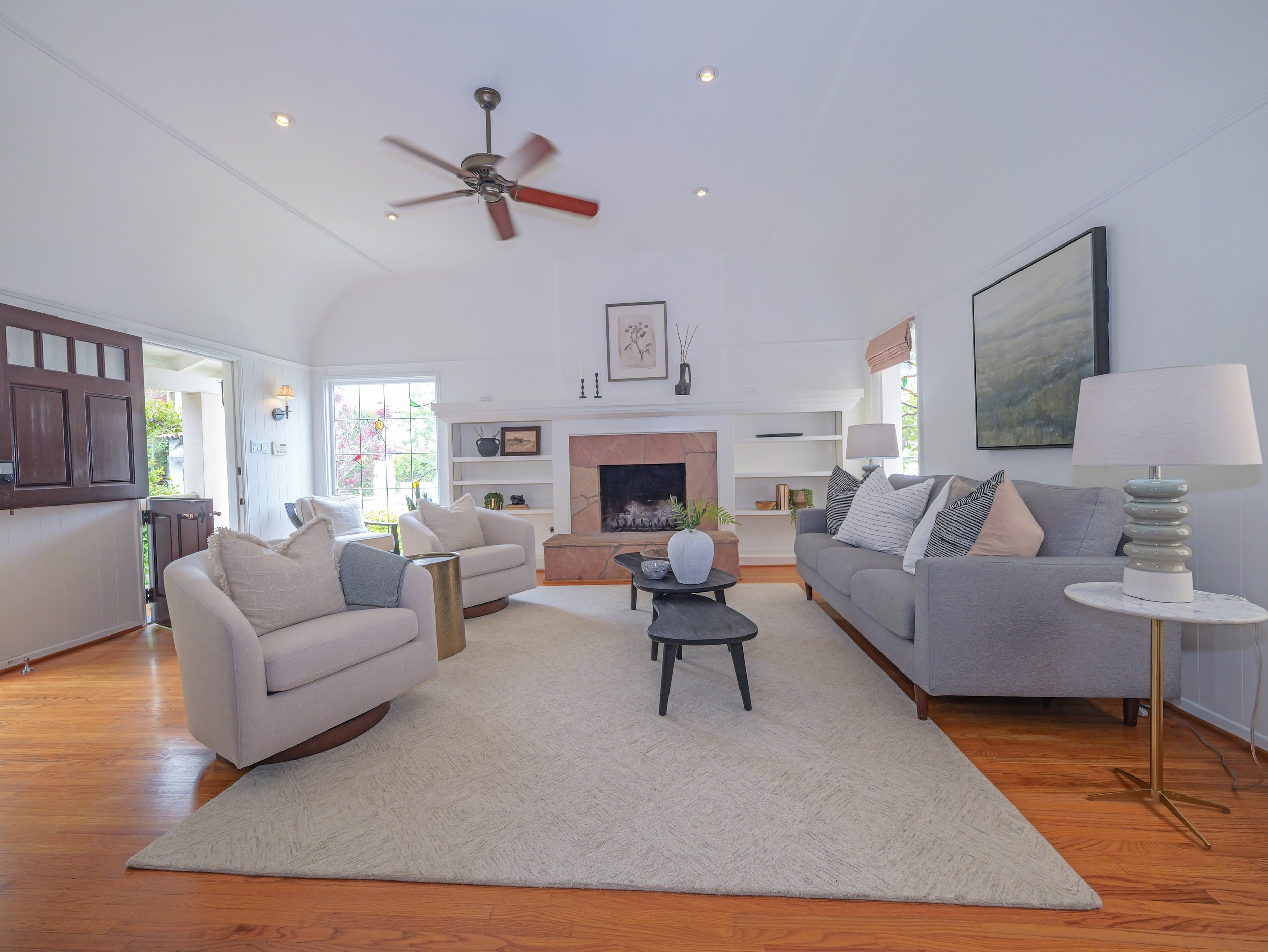 Living room with white walls, a fireplace, beige armchairs, a gray sofa with striped pillows, a wooden side table, a painting above the fireplace, a large wall art piece, and a floor lamp.